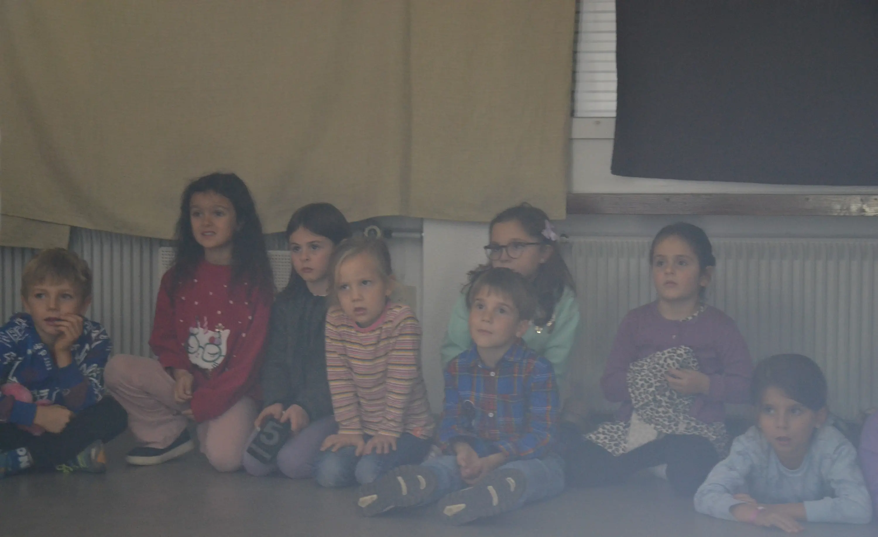 Gruppo di bambini seduti a terra, mentre guardano con attenzione. Alcuni indossano maglioni colorati e si trovano in un ambiente interno con pareti chiare.