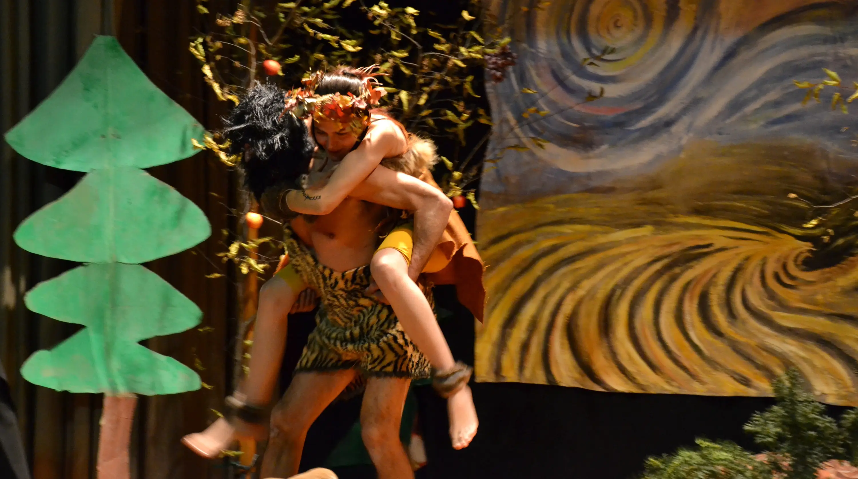 Due personaggi abbigliati in modo primitivo si esibiscono su un palco decorato con alberi e sfondi colorati. Un personaggio maschile porta in spalla un personaggio femminile, entrambi sembrano coinvolti in una danza o una rappresentazione teatr