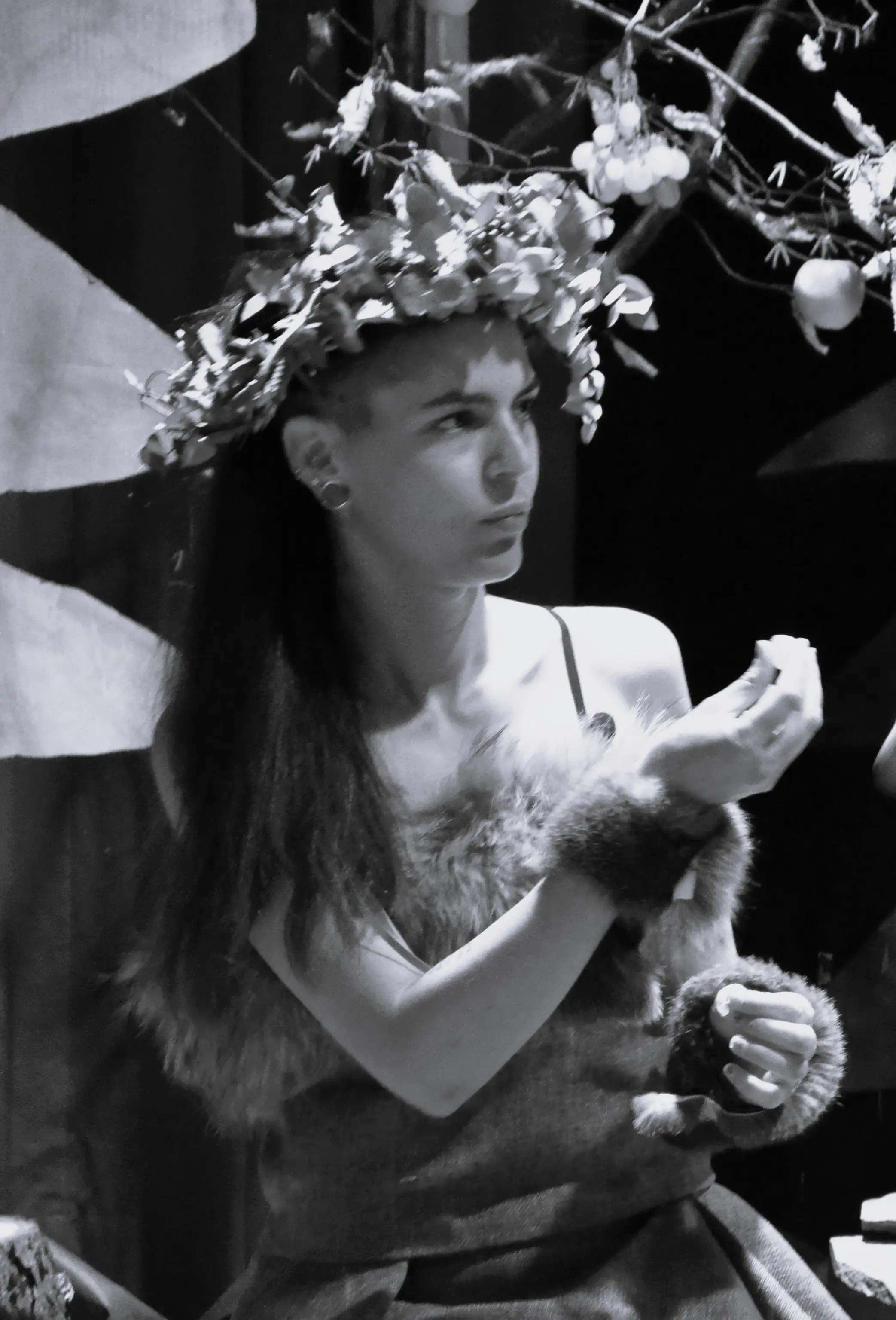 Una giovane donna con una corona di foglie e fiori guarda pensierosa, indossando un abito decorato con pelliccia. Dietro di lei, si intravedono elementi naturali e sfondi artistici.