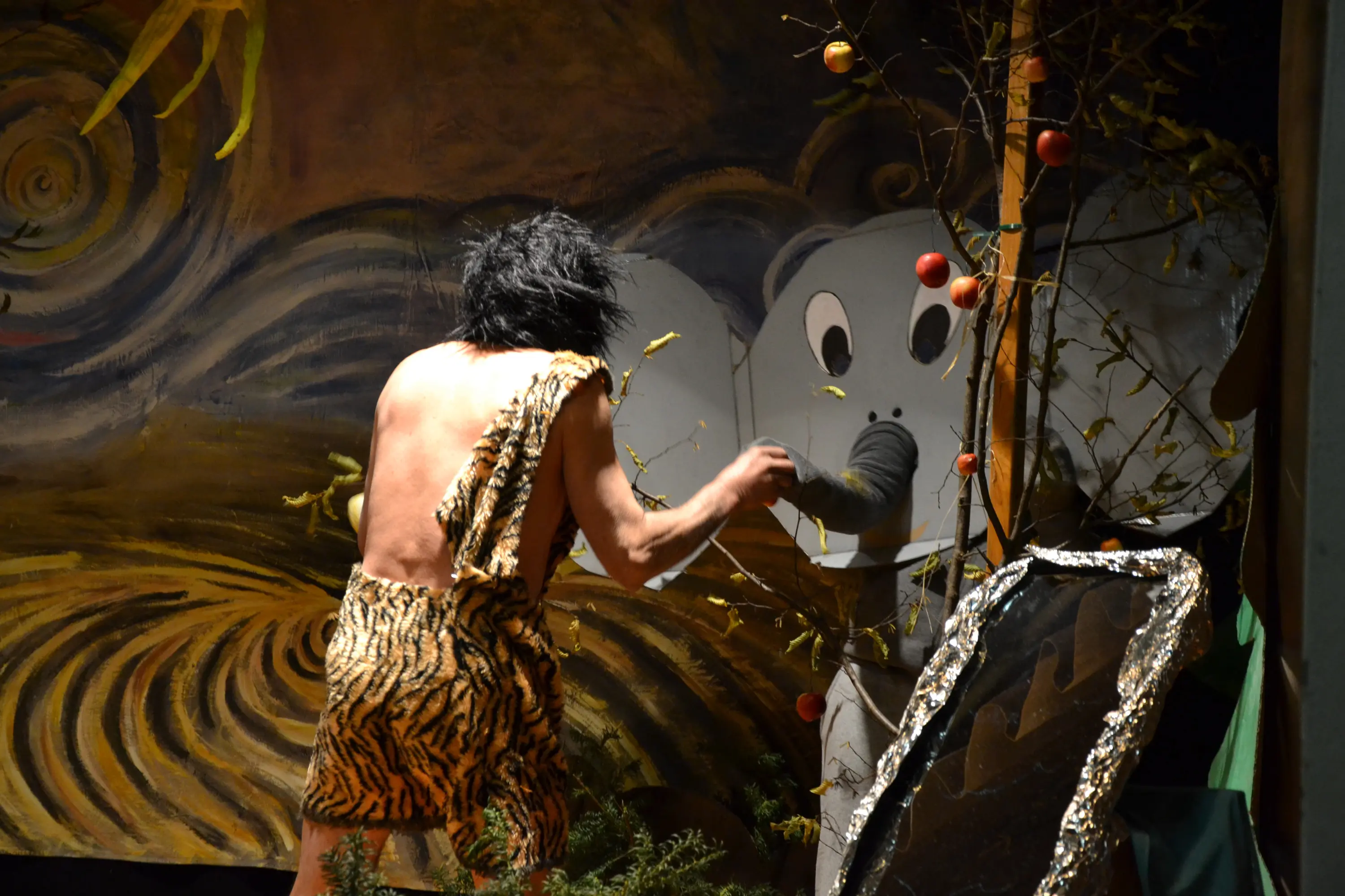 Un uomo vestito con una pelliccia di tigre interagisce con un'illustrazione di un elefante su uno sfondo artistico. Dietro di lui si intravedono alberi e decorazioni naturali.