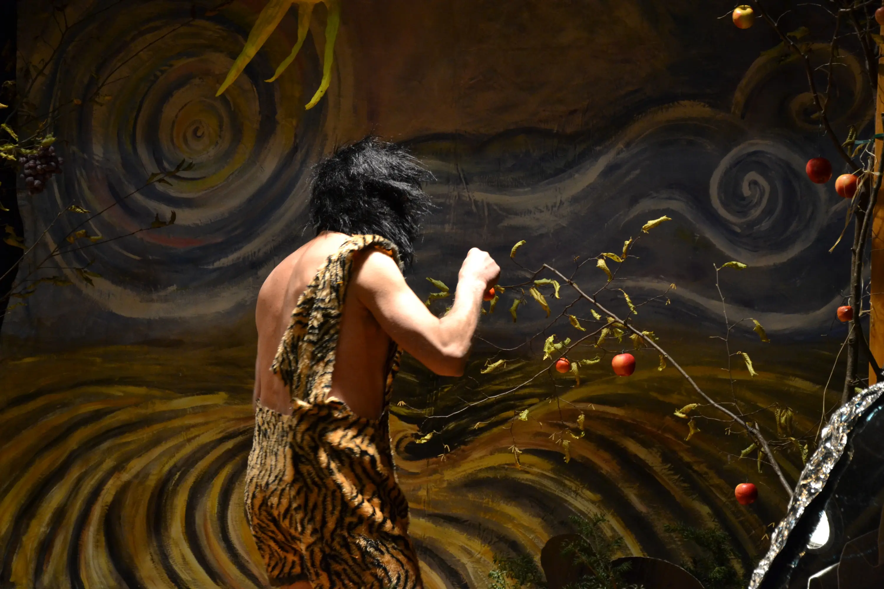 Uomo preistorico con pelli di animali, di spalle, mentre raccoglie mele da un albero in un ambiente dipinto che rappresenta un paesaggio ondulato.