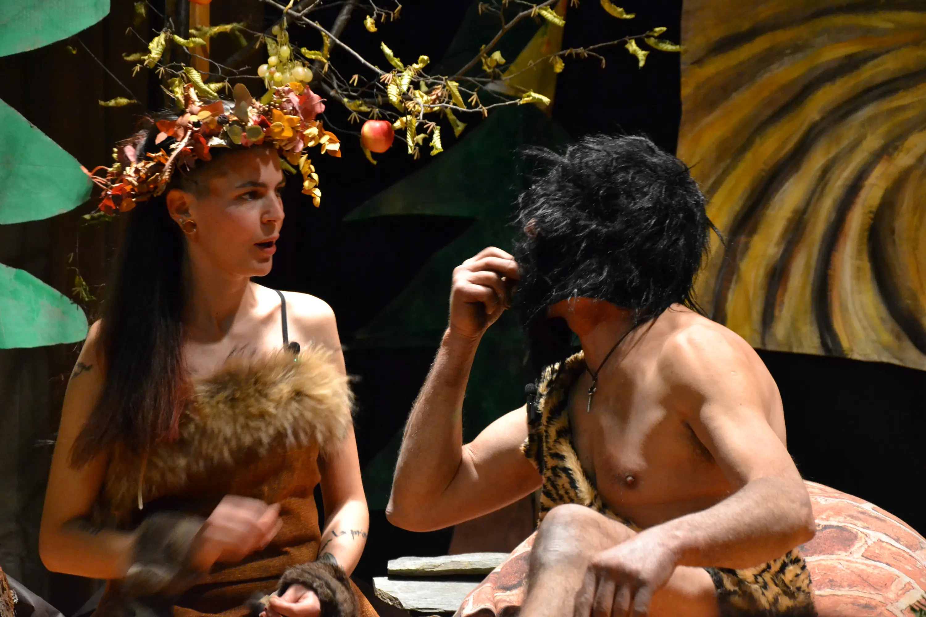 Una giovane donna con una corona di foglie e un vestito di pelliccia conversa con un uomo barbuto, vestito con pelli. L'ambiente è caratterizzato da decorazioni naturali, come un albero con frutti.