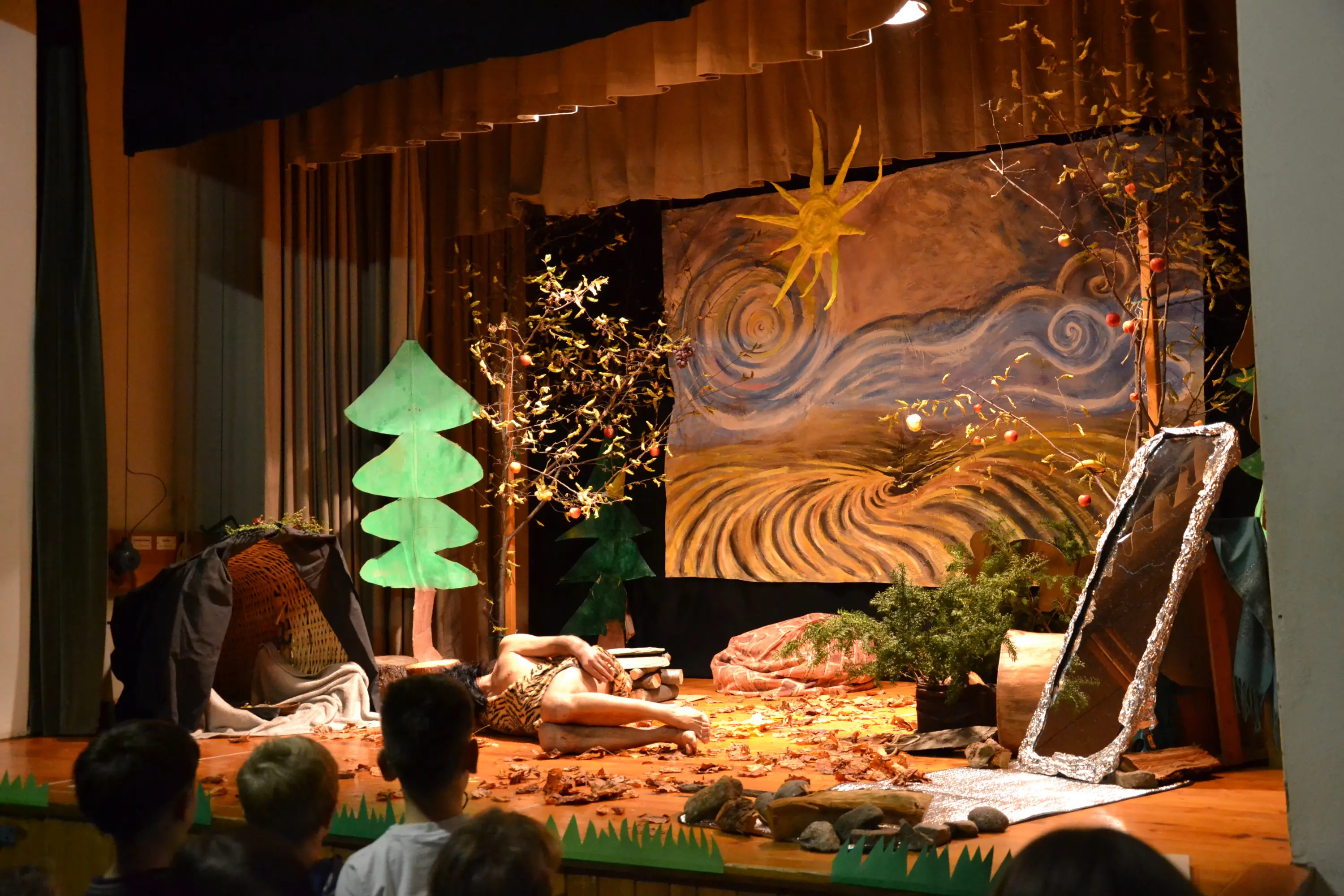 Scena teatrale decorata con alberi di cartone e foglie. Sullo sfondo c'è un grande dipinto con un sole e un paesaggio. Un attore, immerso tra foglie secche, è in posa a terra. Pubblico visibile in basso