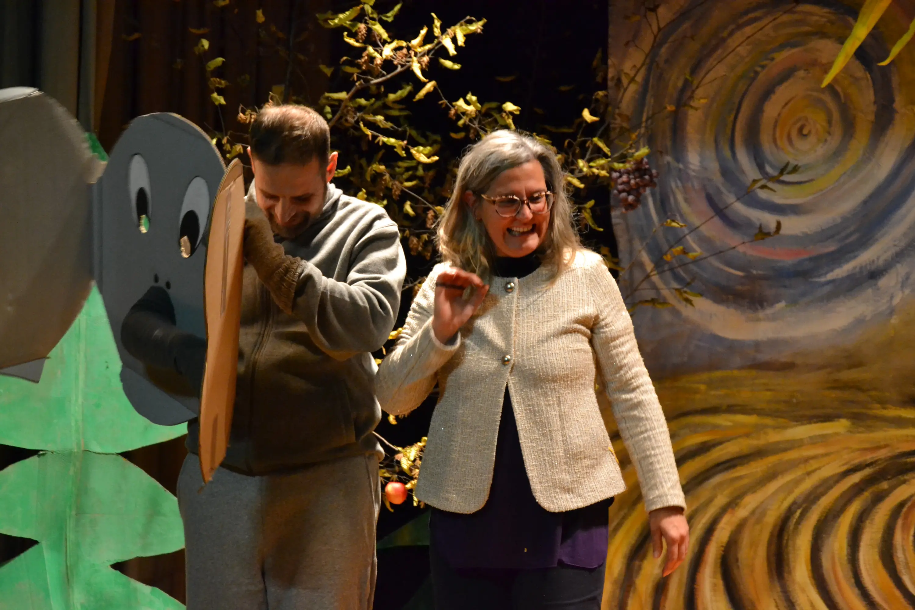 Un uomo indossa una maschera di elefante mentre una donna sorride accanto a lui. Sullo sfondo, un'ambientazione teatrale colorata con elementi naturali.