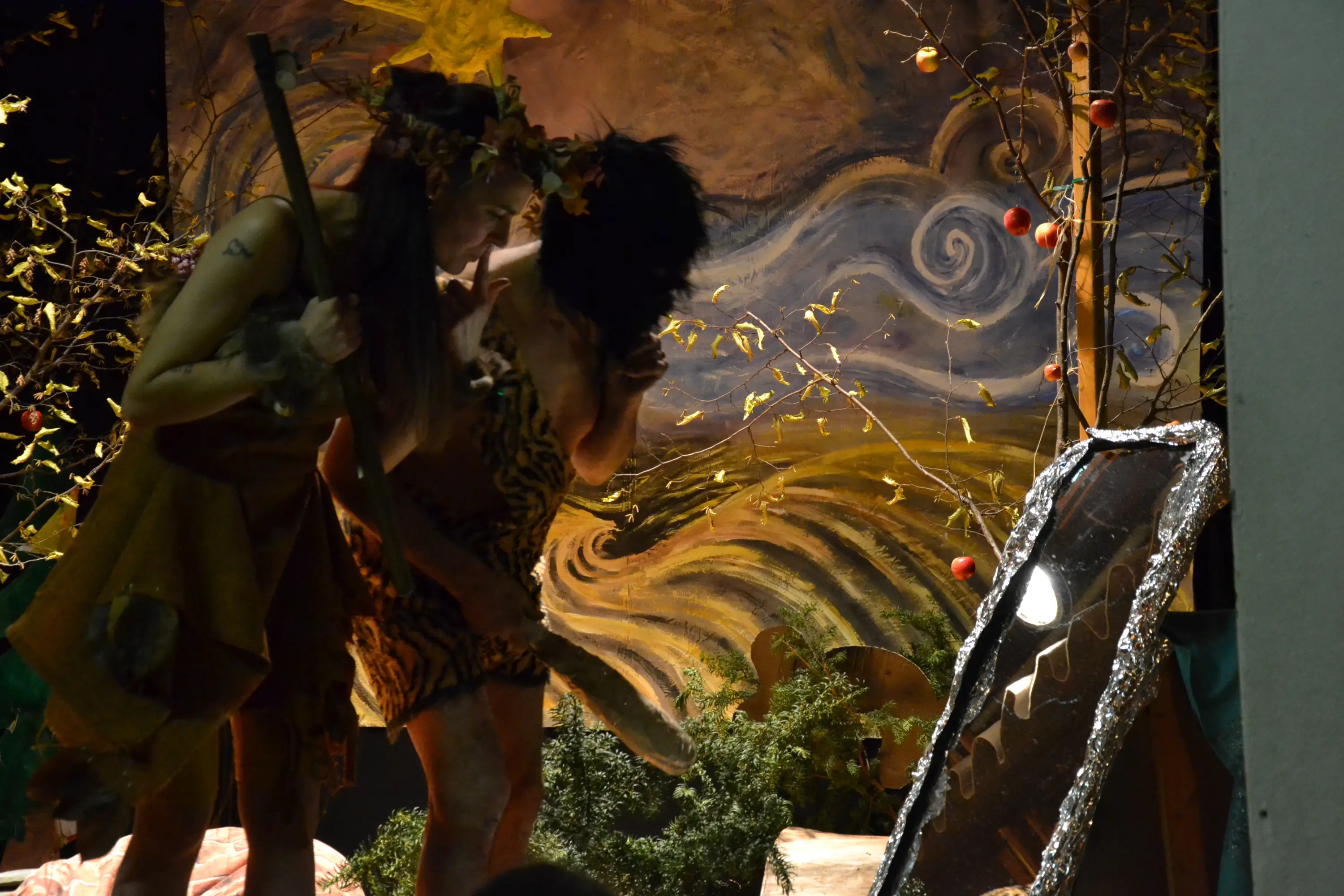 Due persone sul palco, vestite in costumi primitivi, interagiscono davanti a uno specchio. Sullo sfondo, un murale rappresenta un paesaggio naturale con onde e vegetazione. Atmosfera teatrale e suggestiva.