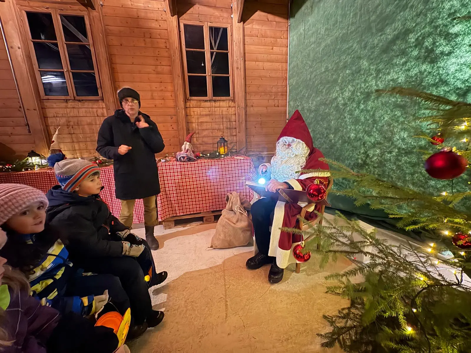 Babbo Natale seduto con un sacco di regali, circondato da un albero di Natale decorato. Due bambini lo osservano con attenzione mentre una donna adulta parla. Lo sf