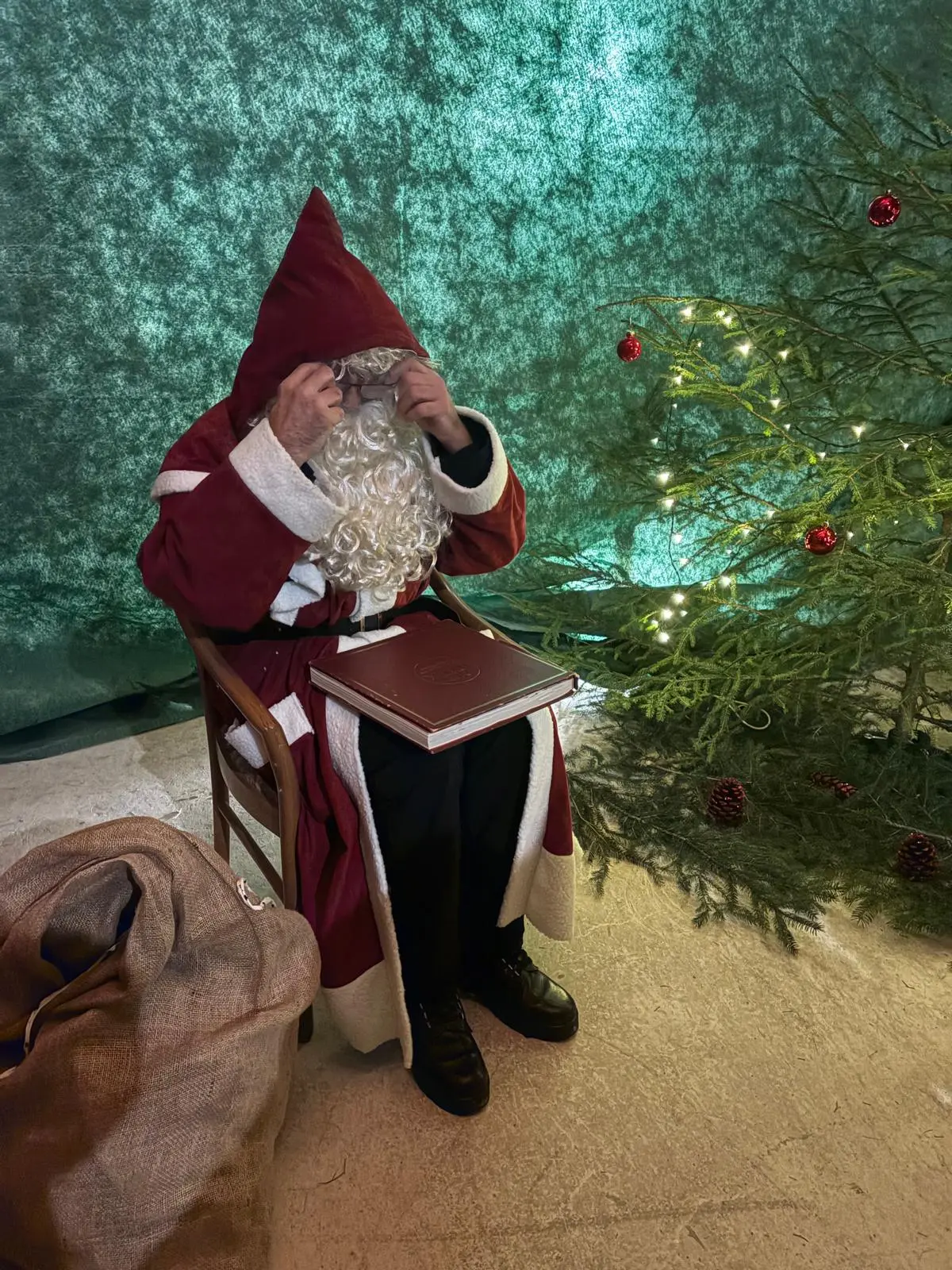 Babbo Natale seduto su una sedia, con un grande libro in grembo, mentre indossa un cappuccio rosso. Dietro di lui, un albero di Natale decorato