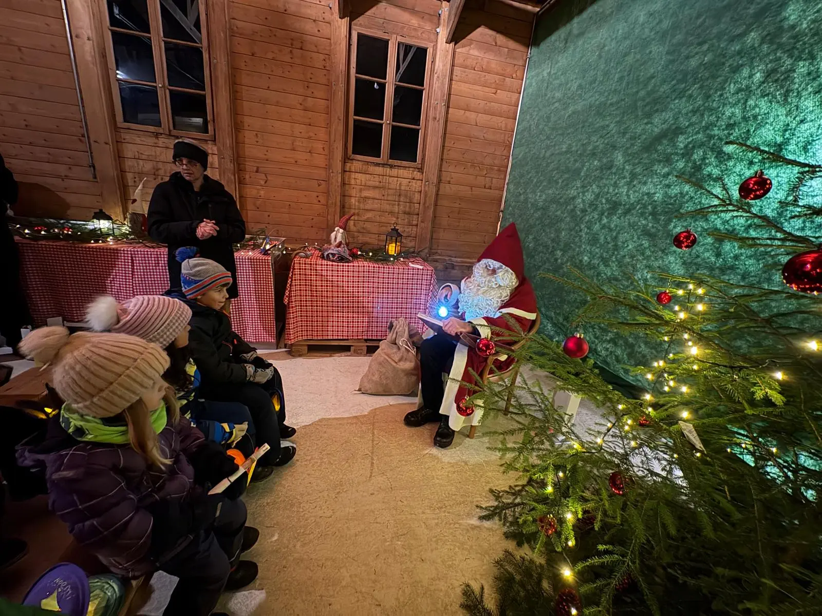 Babbo Natale seduto su una sedia all'interno di un rifugio di legno, circondato da luci natalizie e decorazioni. Un gruppo di bambini, vestiti