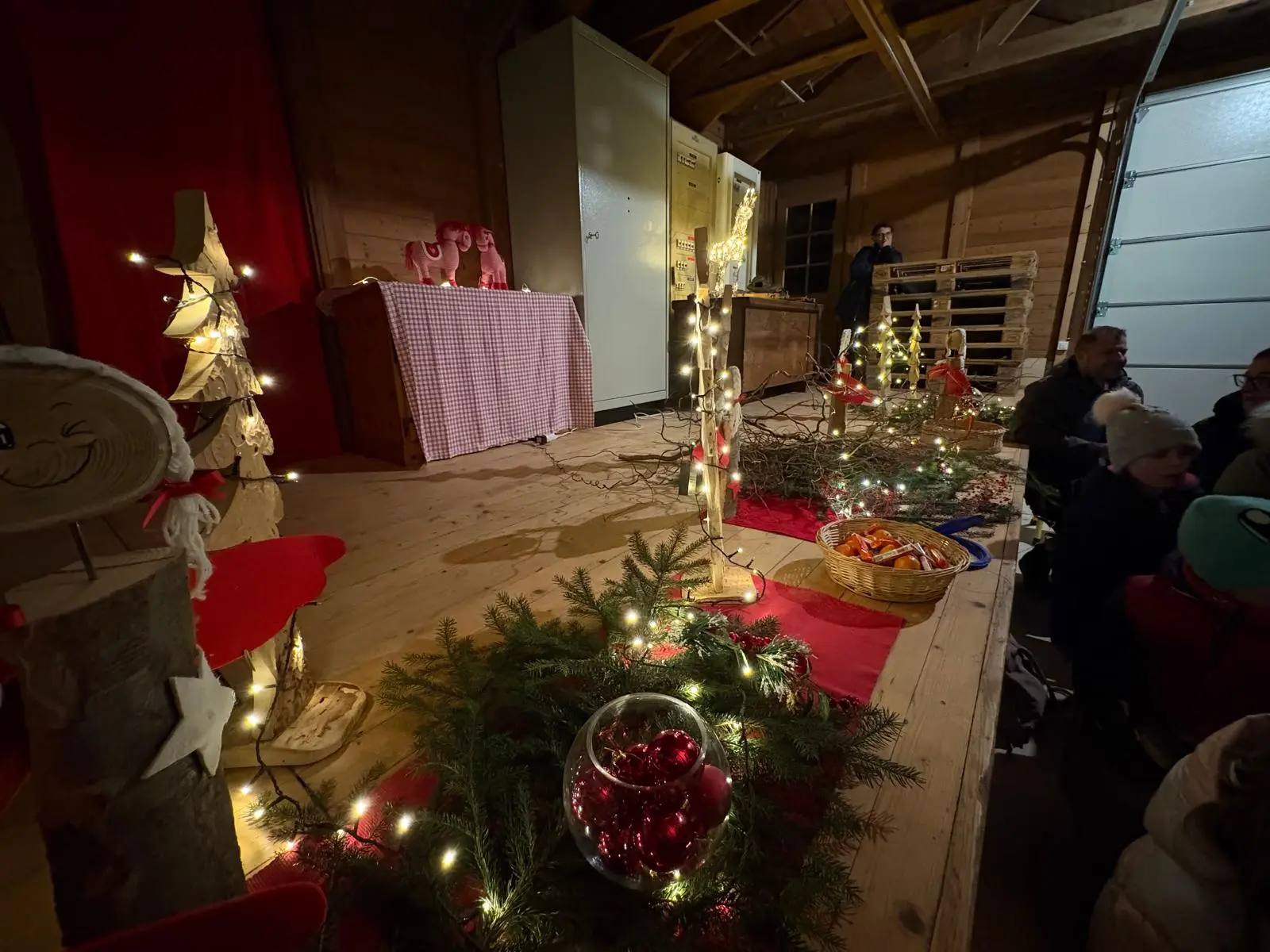 Un palco di legno decorato per le festività, con un tavolo addobbato, luci natalizie e decorazioni come alberi di legno e piante verdi. Alcuni