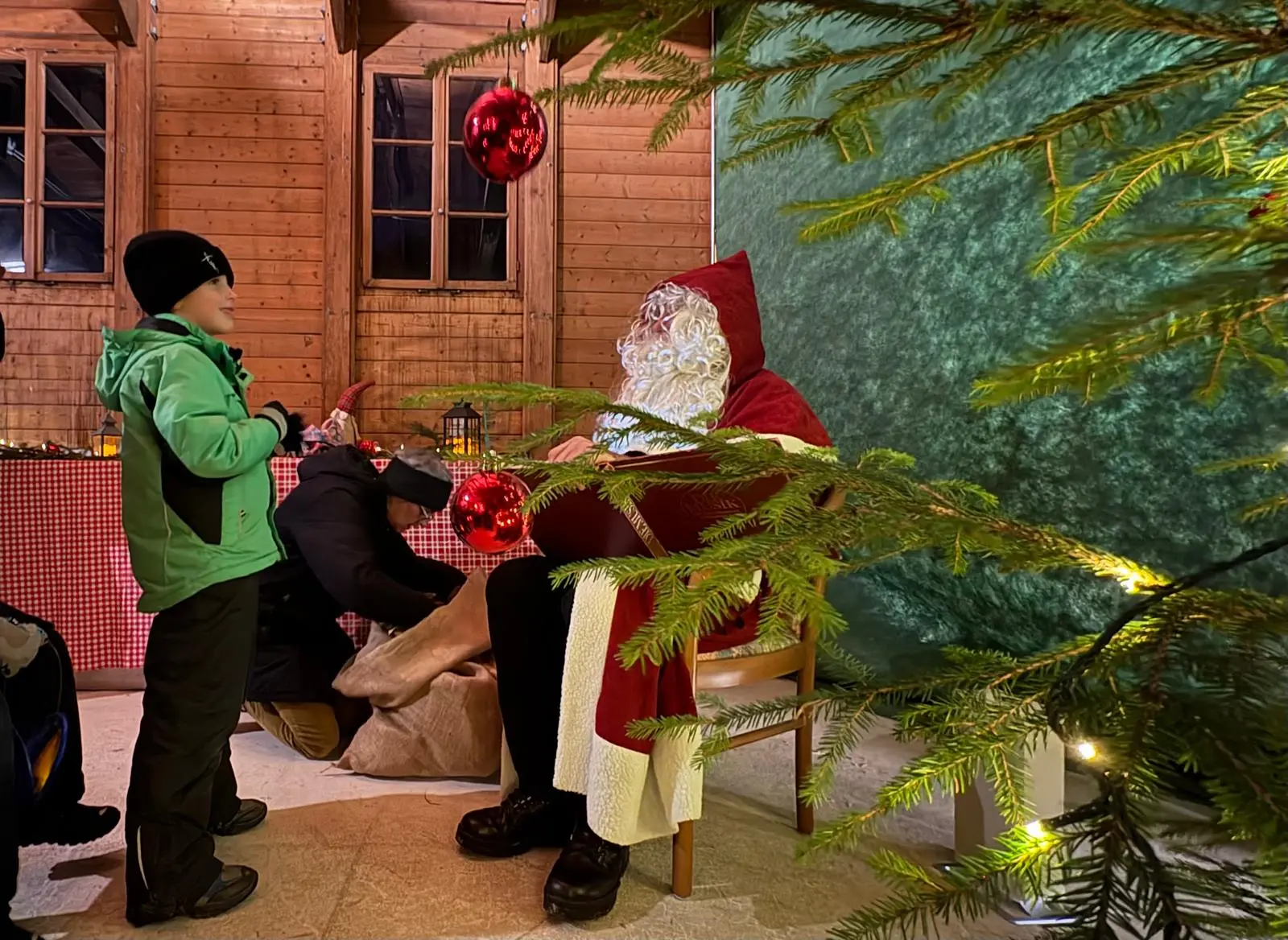 Un bambino in giacca verde parla con Babbo Natale, seduto su una sedia, con una lunga barba bianca. Sullo sfondo, un albero di Natale decorato con pall