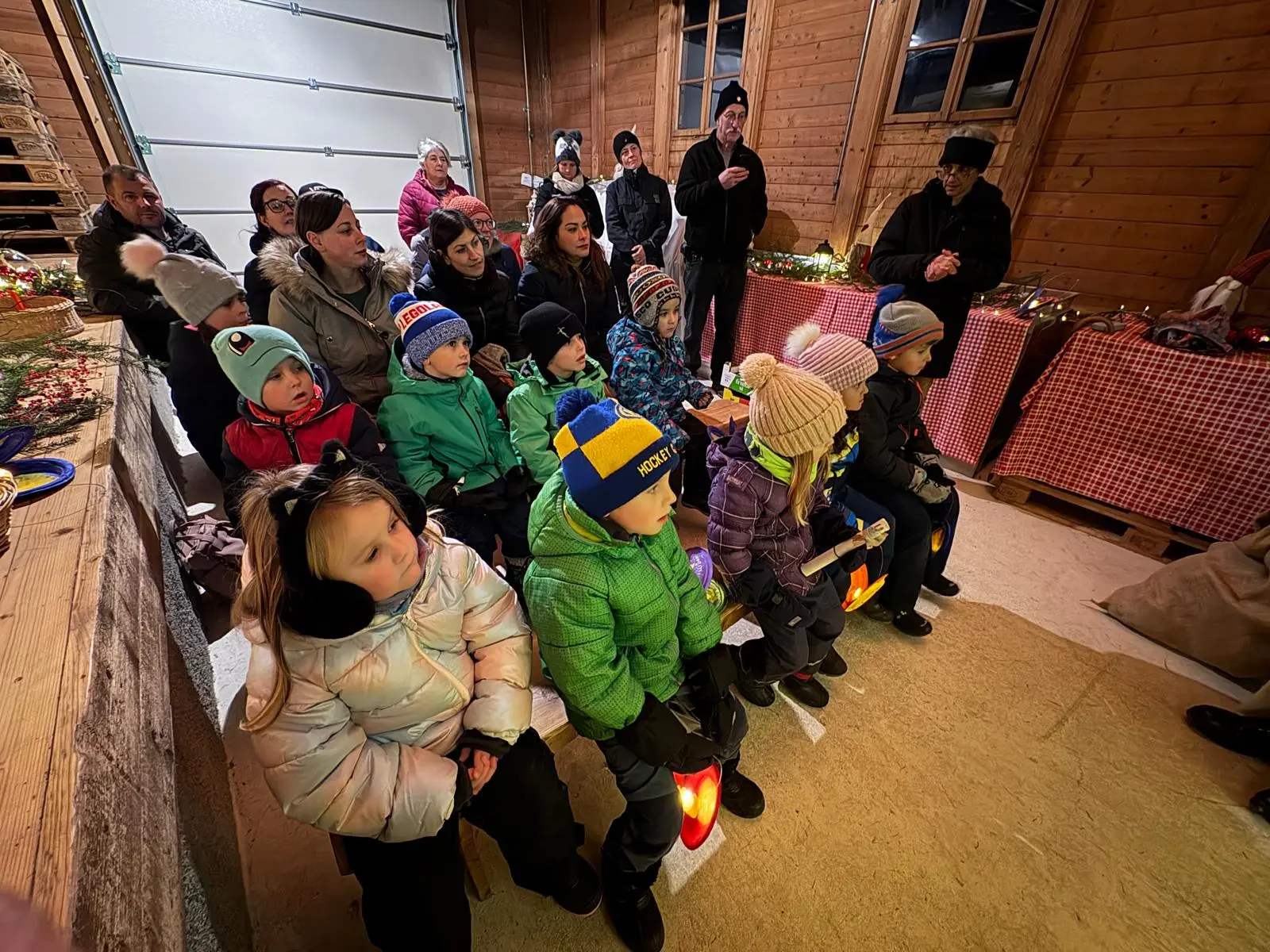 Un gruppo di adulti e bambini seduti in una stalla di legno decorata per un evento festivo. I bambini indossano giacche colorate e cappelli, mentre gli