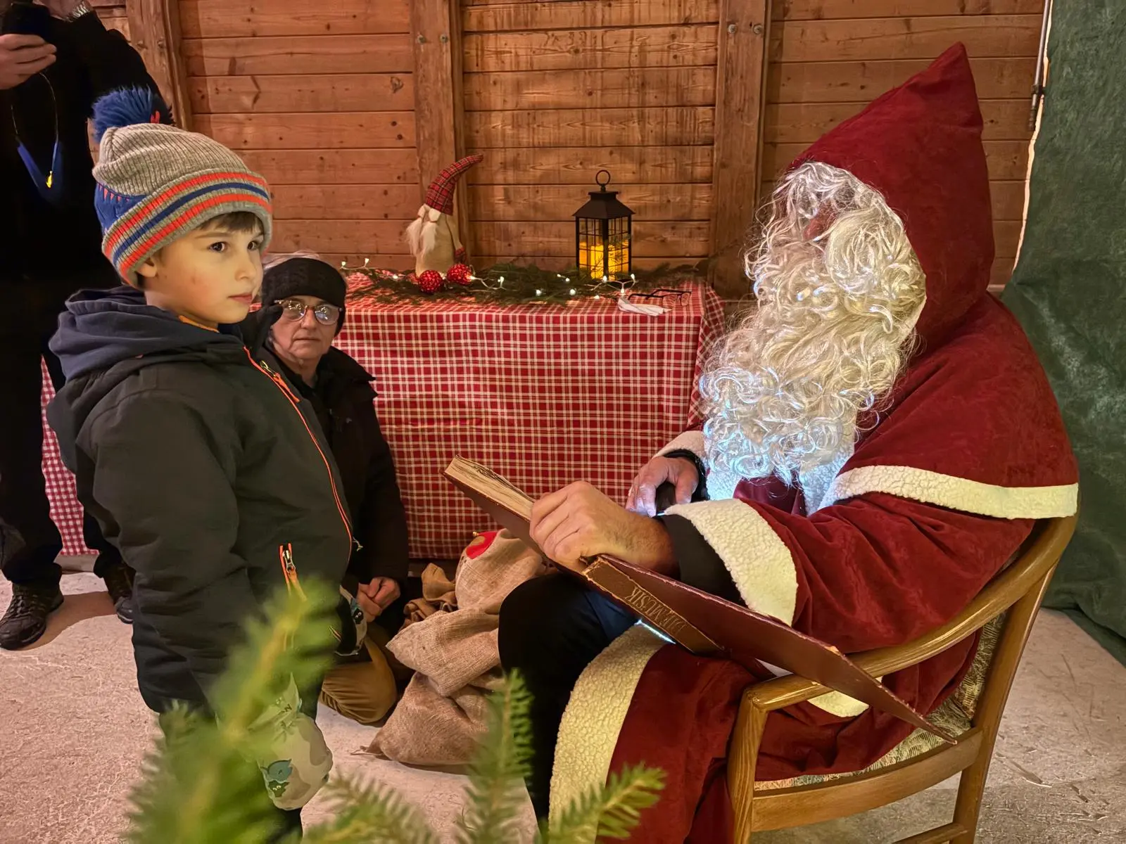 Un bambino di fronte a Babbo Natale, che indossa un grande costume rosso e ha una lunga barba bianca. Sullo sfondo, un tavolo decorato con una tovaglia a