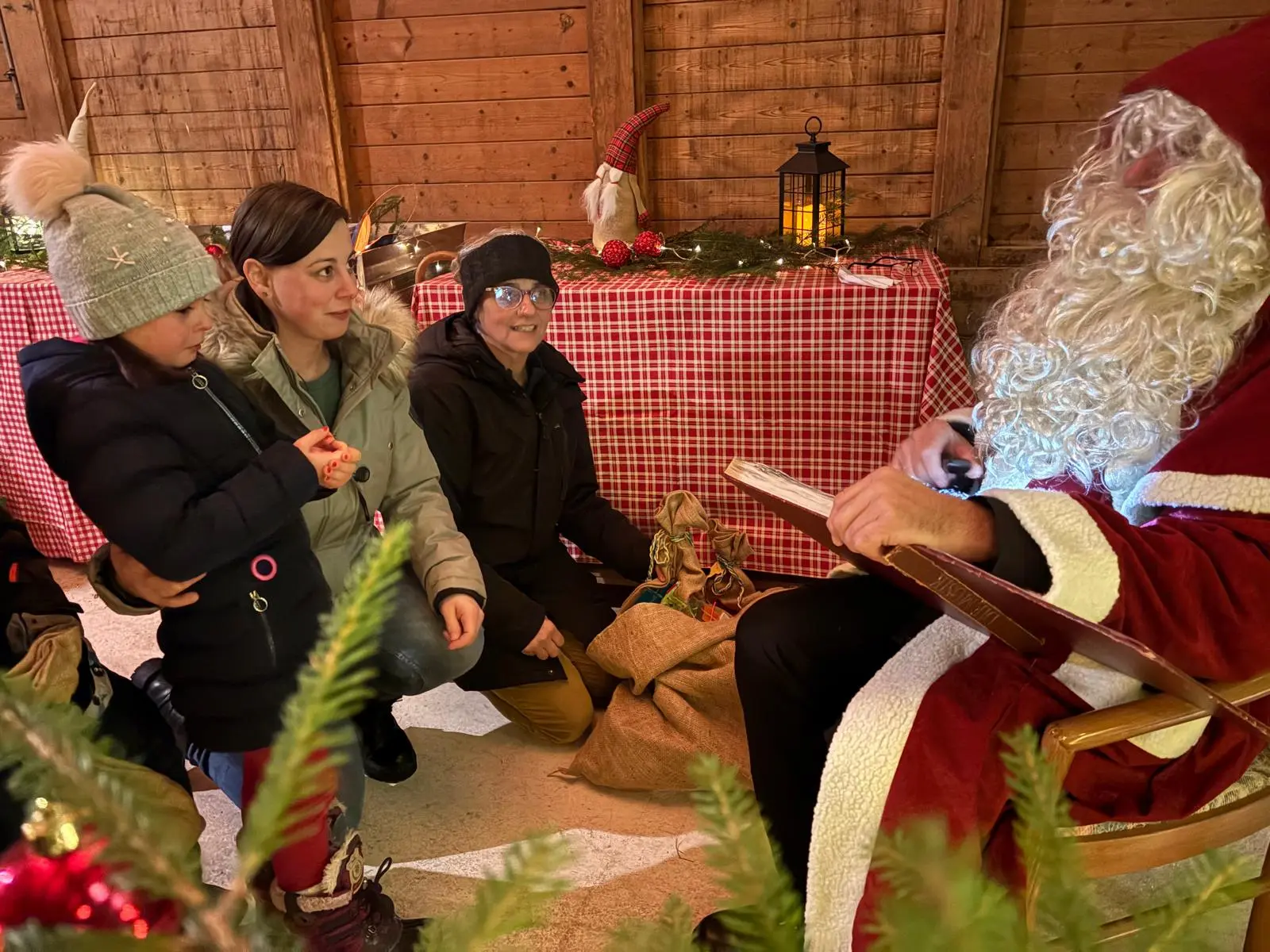 Una donna con un cappotto grigio e un cappello invernale guarda attentamente Babbo Natale, seduto su una sedia, mentre tiene un libro. Una bambina accanto