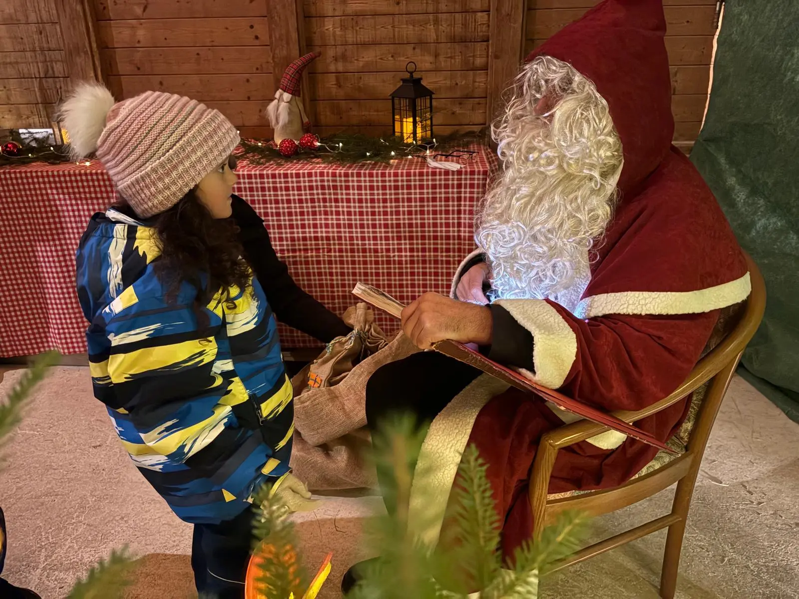 Un bambino con un giubbotto blu e giallo parla a Babbo Natale, che indossa un abito rosso e una lunga barba bianca. Dietro di loro si vede