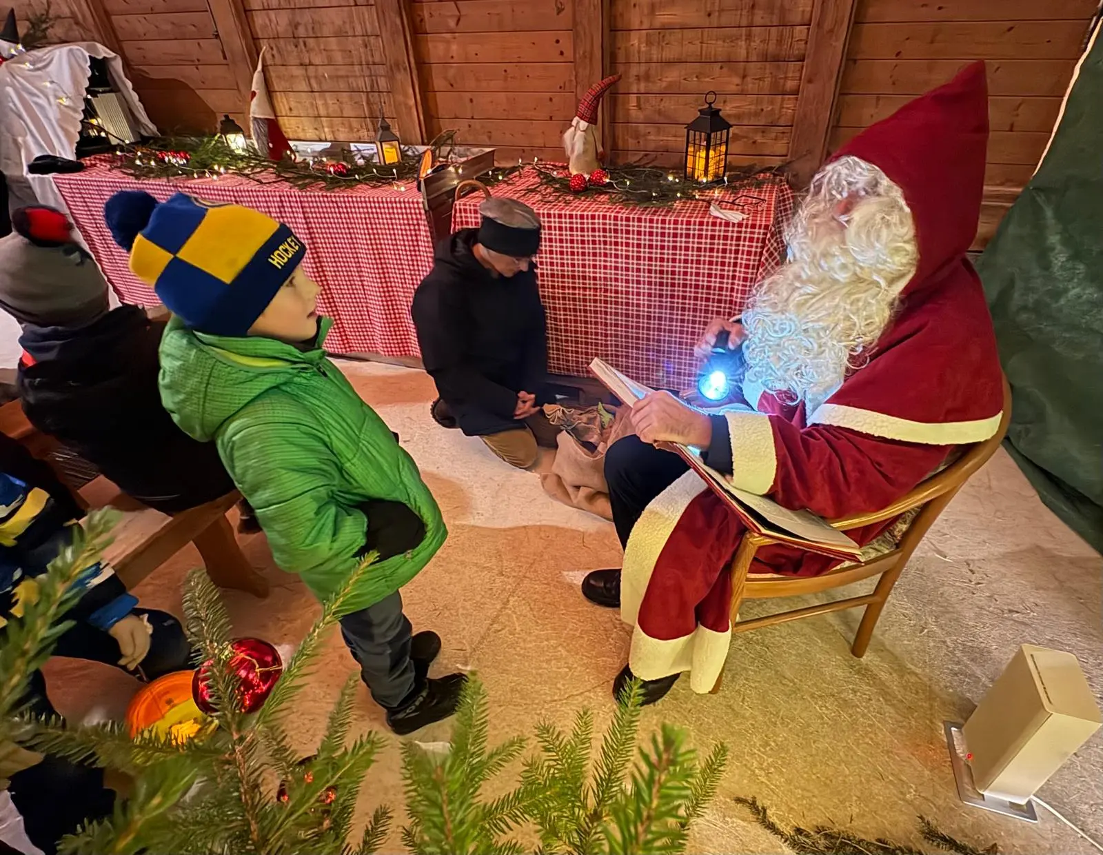 Babbo Natale con una lunga barba bianca è seduto su una sedia, mentre legge un libro illuminato da una torcia. Un bambino in giacca verde e cappello a strisce