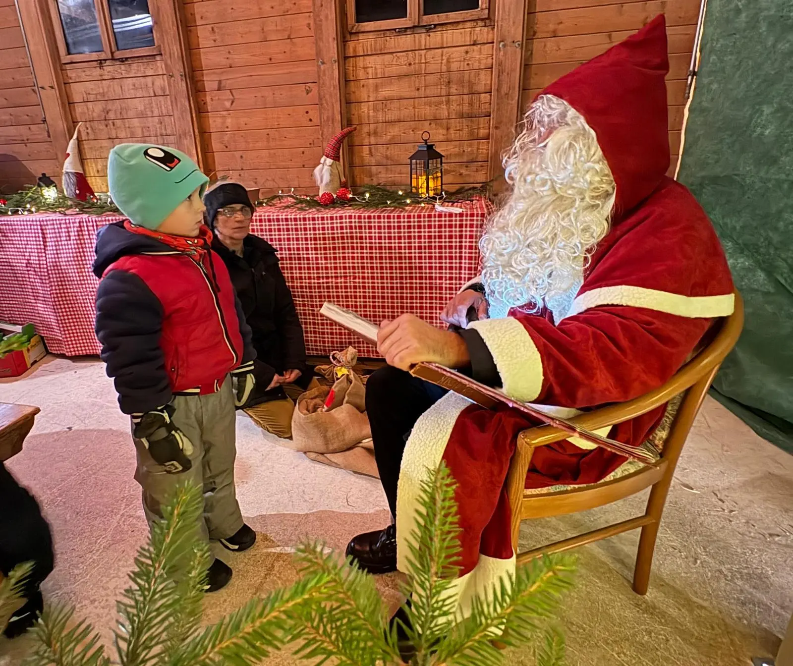 Un bambino in giacca rossa ascolta attentamente Babbo Natale, vestito con un mantello rosso e barba bianca, seduto su una sedia in un ambiente festivo decor