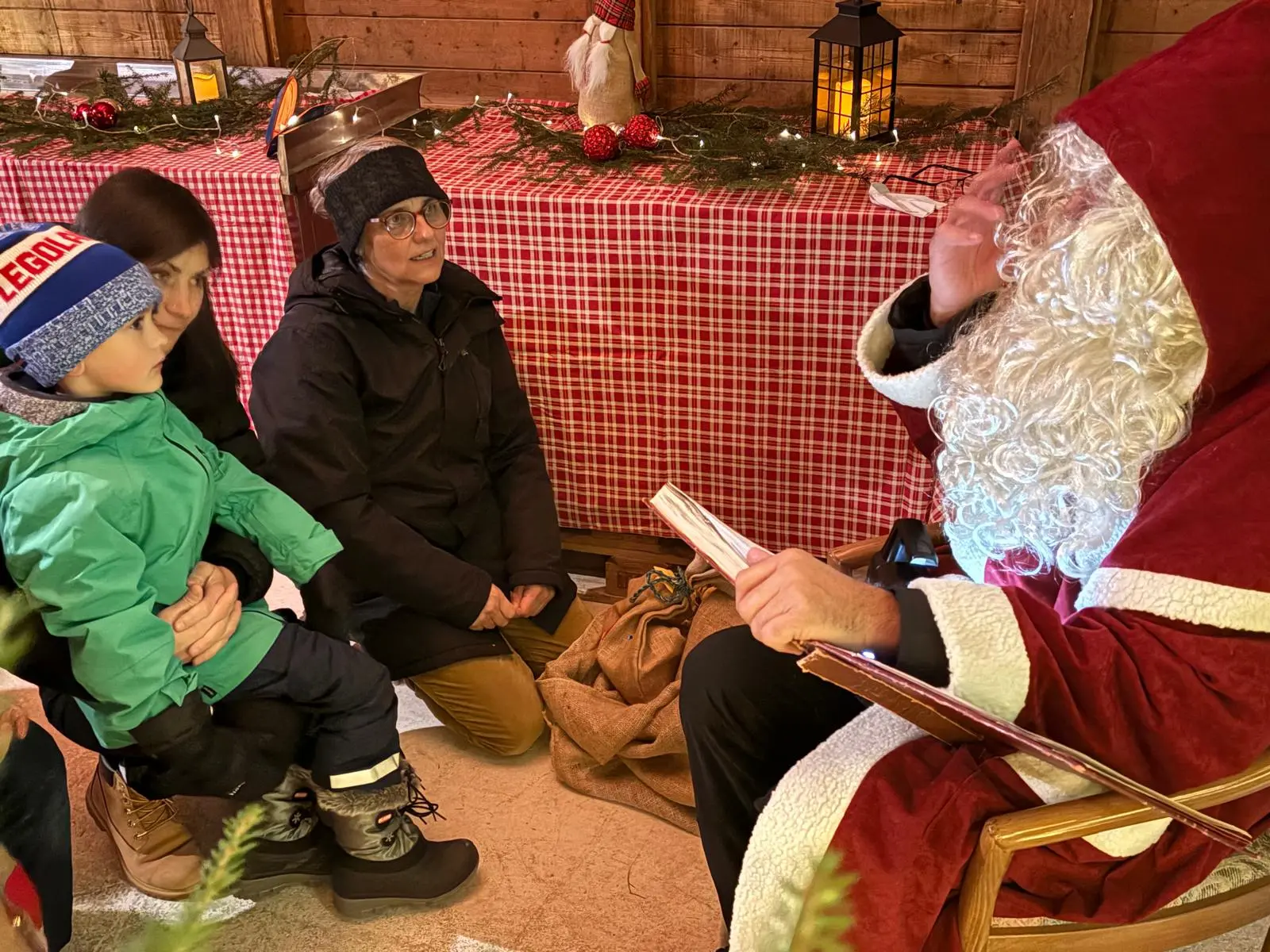 Babbo Natale seduto su una sedia in un ambiente natalizio, mentre ascolta e parla con una madre e il suo bambino, vestito con una giacca verde e un berretto blu