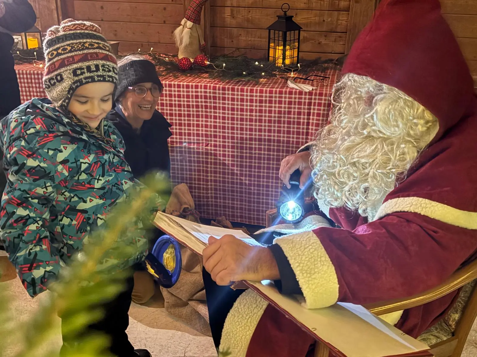 Un bambino sorride mentre guarda Babbo Natale, che legge un libro illuminato da una torcia. Una persona adulta, sorridente, osserva dalla sinistra. Sullo sfondo, decor