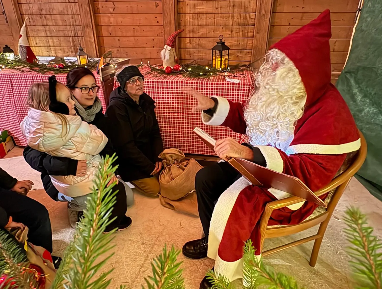 Babbo Natale seduto su una sedia, circondato da decorazioni natalizie, racconta una storia a una famiglia che lo ascolta con attenzione. Una giovane donna tiene in braccio