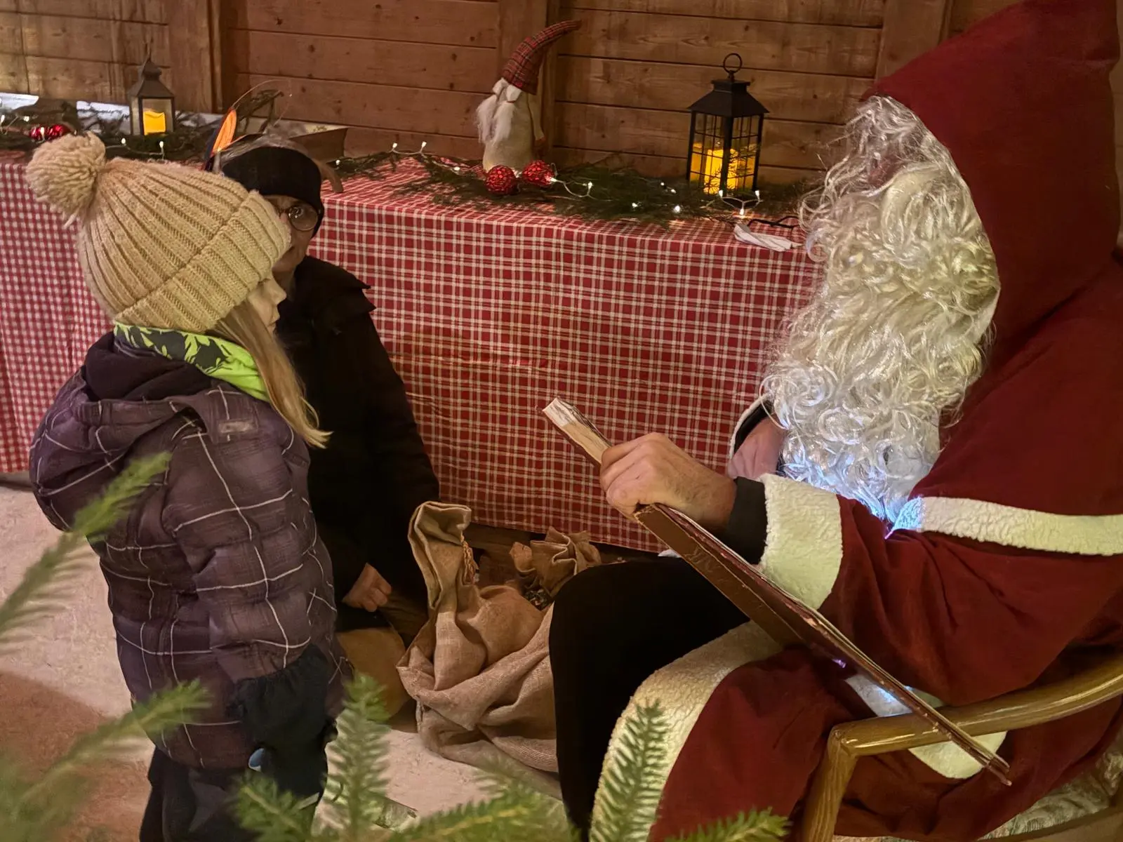 Babbo Natale, seduto in un'atmosfera festosa, legge una lista mentre un bambino lo ascolta attentamente. Una persona adulta è presente in background. Decorazioni natalizie e lan