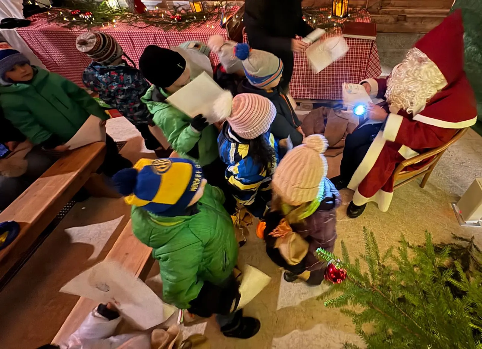 Un gruppo di bambini circonda Babbo Natale, che è seduto e sorride. I bambini indossano cappelli e giacche invernali, mentre mostrano i loro dis