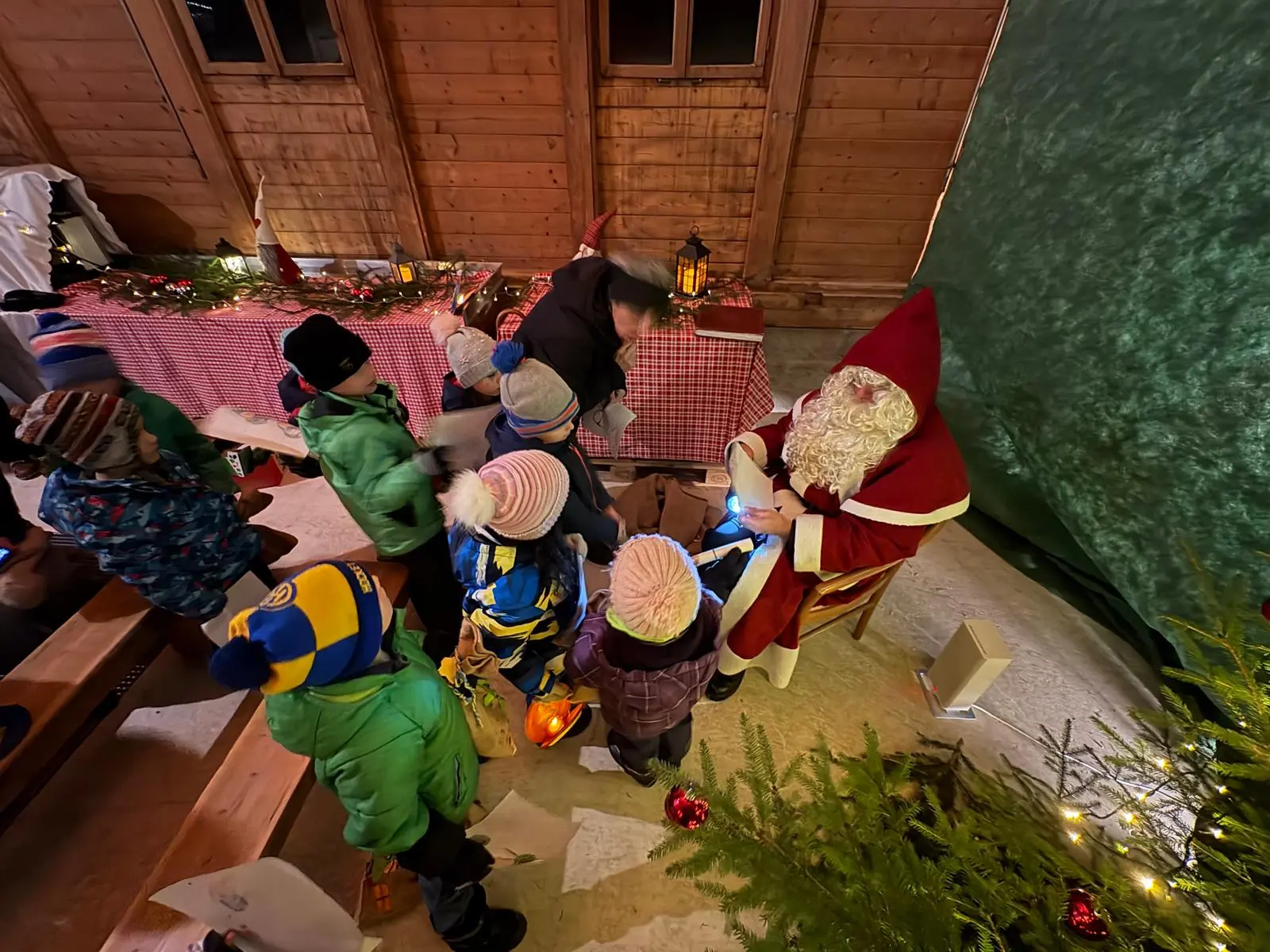 Babbo Natale è seduto su una sedia circondato da bambini, mentre uno di loro si avvicina per mostrargli un disegno. Alle spalle, decorazioni natalizie