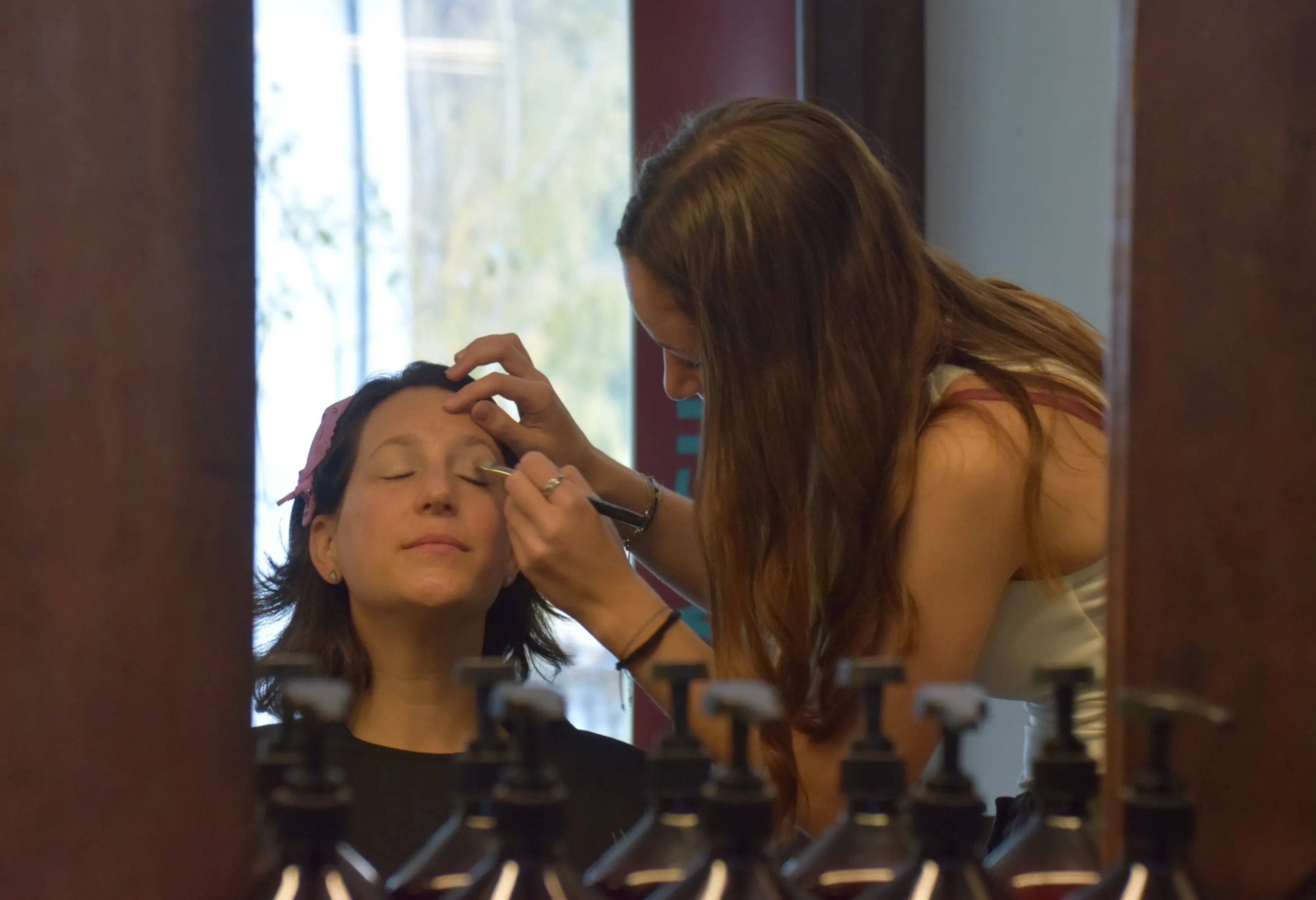 Due donne in un salone di bellezza. Una di esse sta ricevendo un trattamento per le sopracciglia mentre l'altra la assiste. In primo piano, si vedono flaconi di prodotti per la cura della pelle.