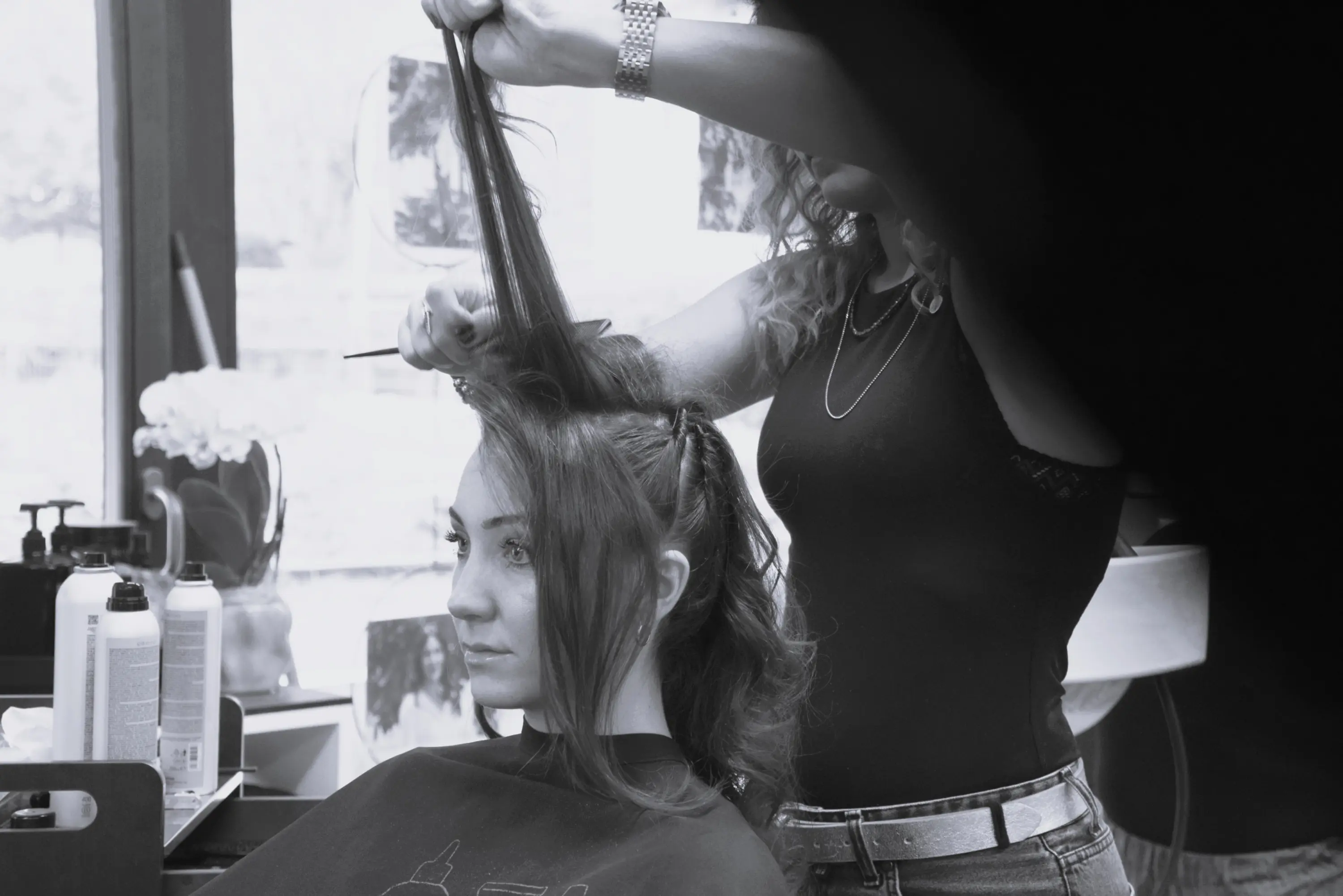Una donna seduta in un salone di bellezza, mentre una parrucchiera le acconcia i capelli. I prodotti per capelli sono visibili sulla postazione.