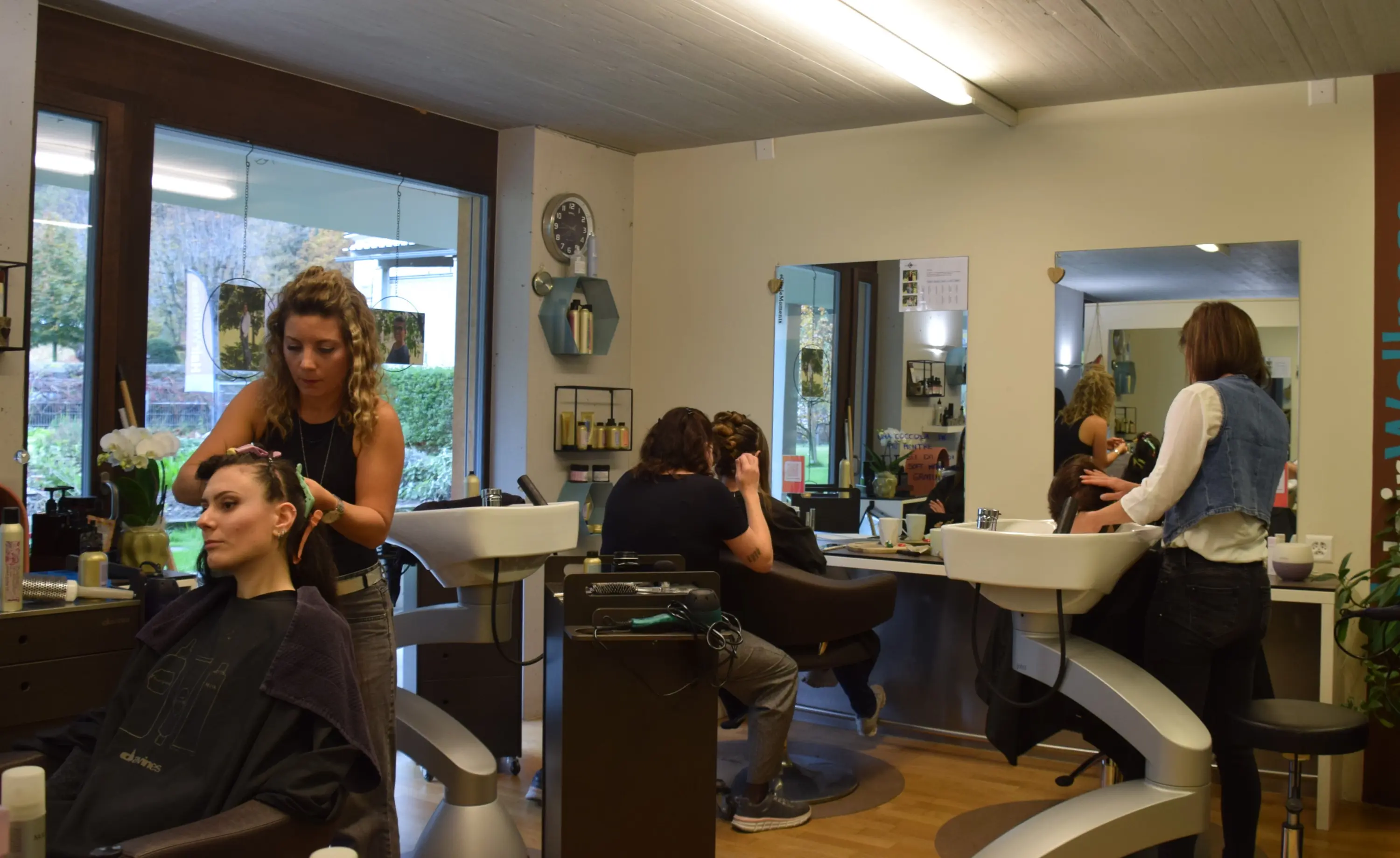 Due parrucchieri al lavoro in un salone. Una donna è seduta su una poltrona mentre riceve un trattamento ai capelli. Un'altra cliente, in piedi, si guarda allo specchio. Nel salone si vedono attrezzature e prodotti per la cura dei capelli.