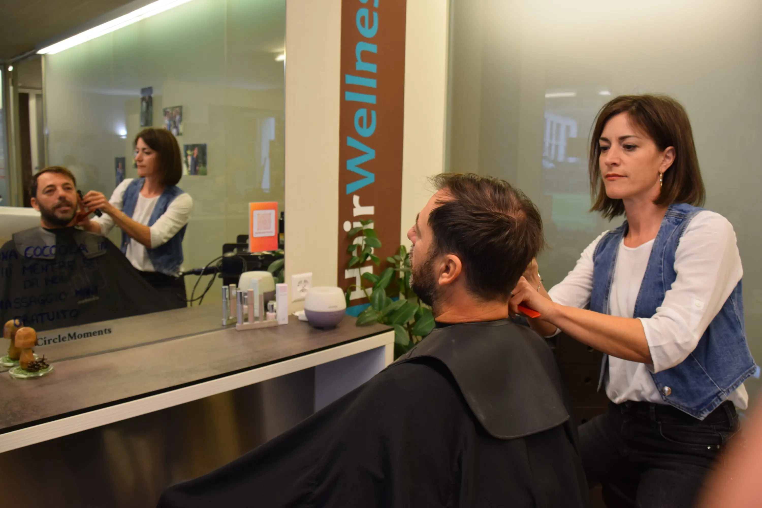 Un parrucchiere sta pettinando un uomo di spalle, riflettendo la sua immagine nello specchio. La parrucchiera è concentrata nel suo lavoro.