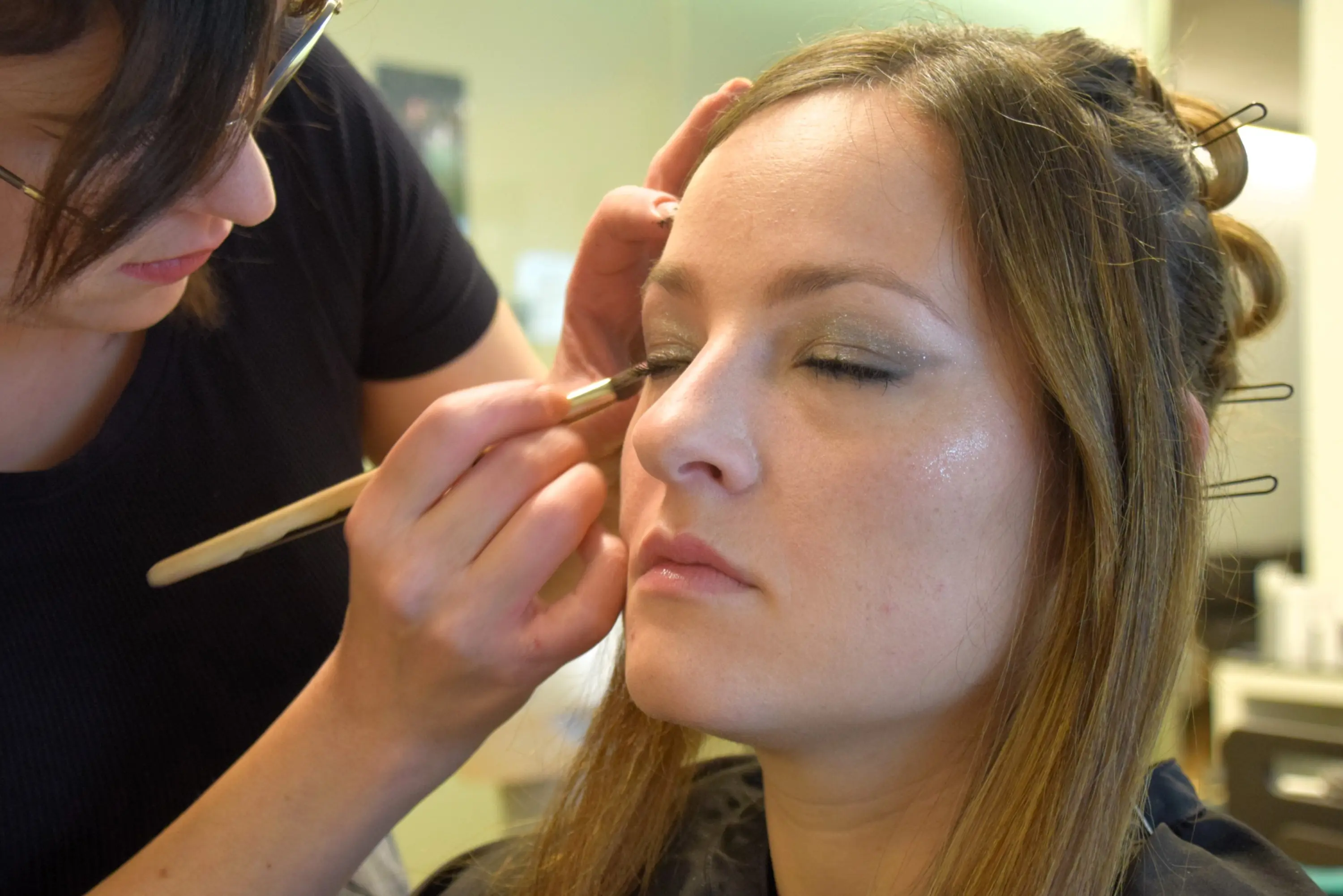 Una truccatrice applica il trucco sugli occhi di una donna seduta, concentrata e con i capelli in parte raccolti.