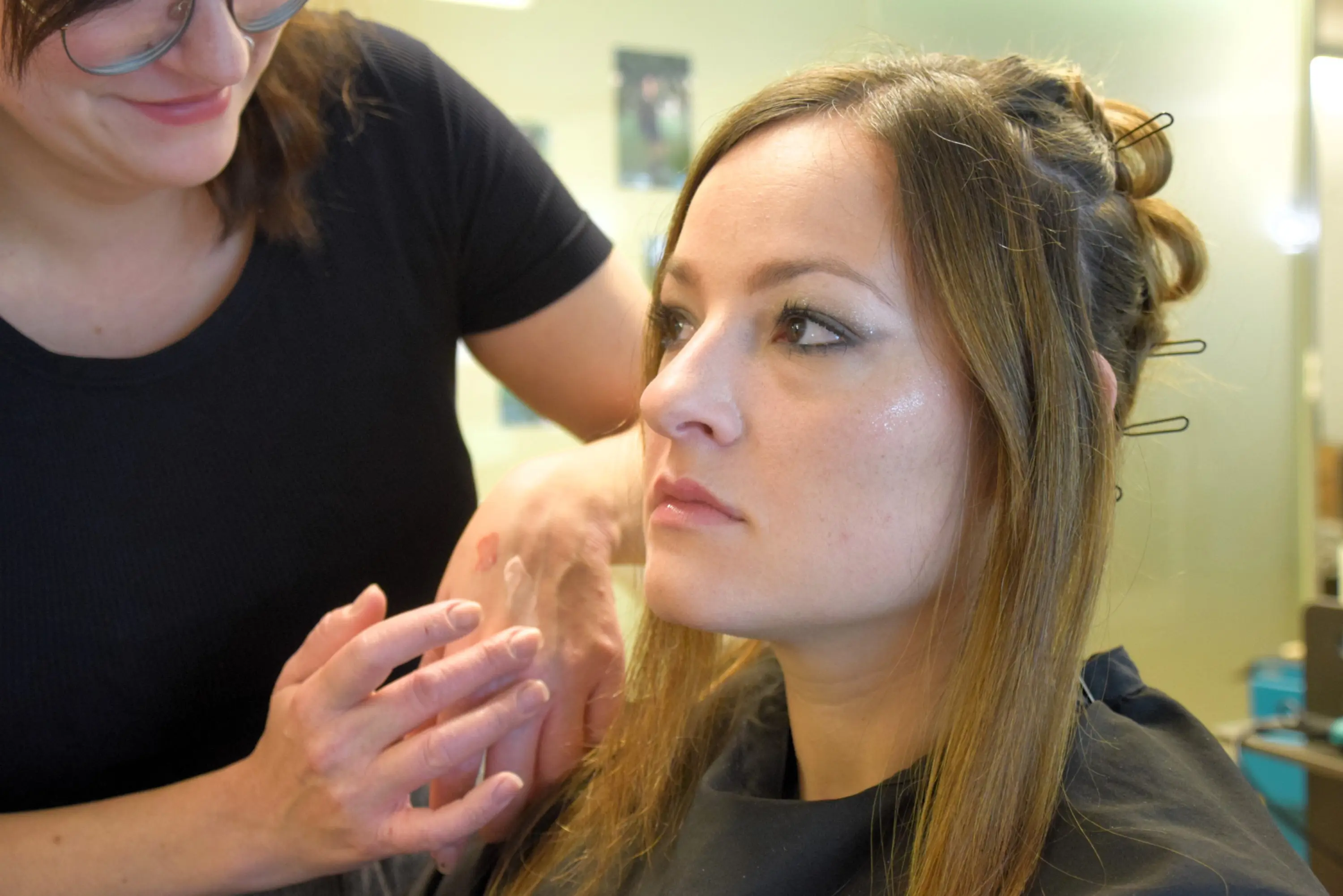 Una donna con capelli lunghi e castani, seduta su una sedia, osserva attentamente mentre un'altra persona le applica del trucco sul viso. L'atmosfera è di preparazione per un evento o una sessione fotografica.