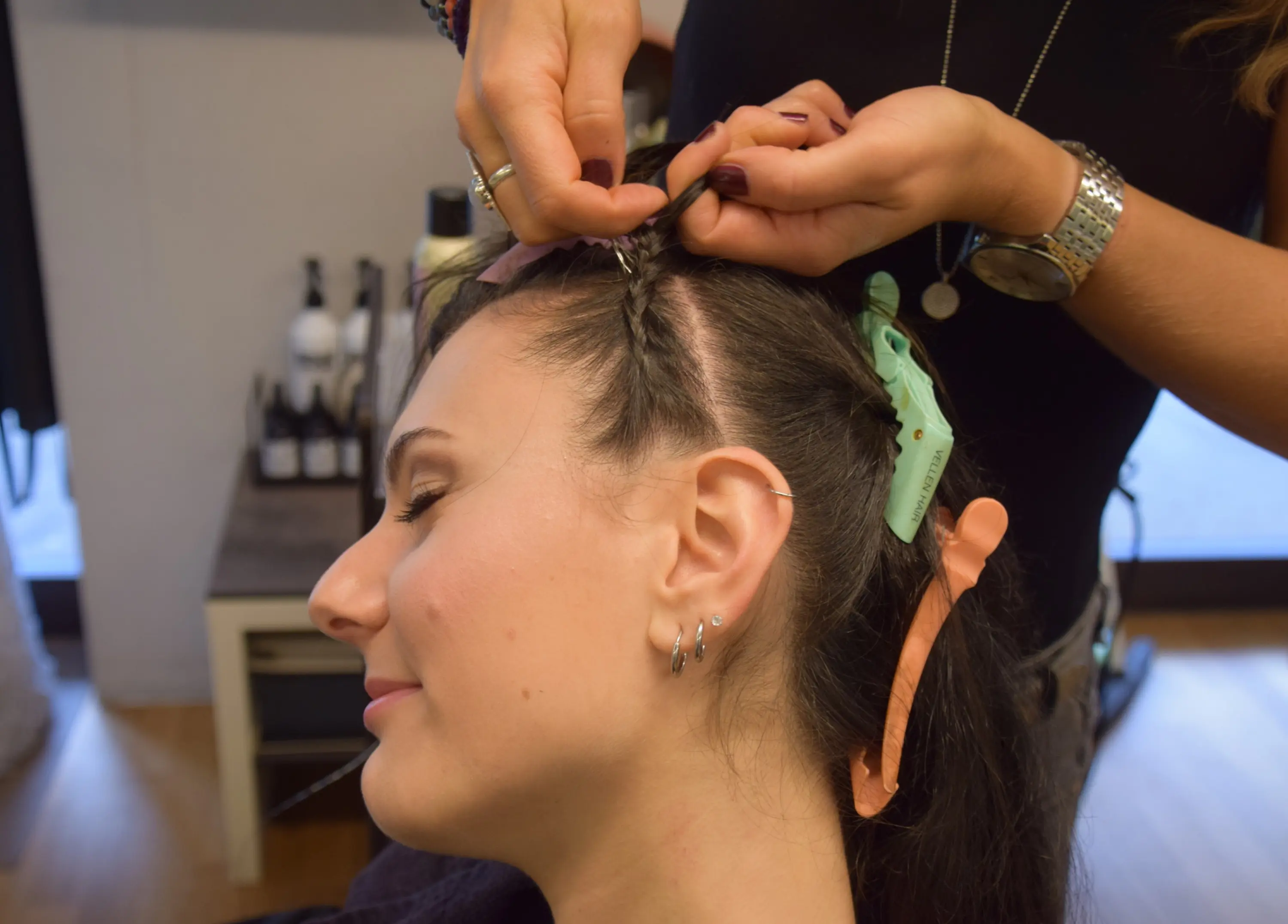 Una donna con i capelli lunghi e scuri sta ricevendo un'acconciatura in un salone. Una stylist le sta intrecciando i capelli, utilizzando fermagli colorati per tenere le ciocche in posizione.
