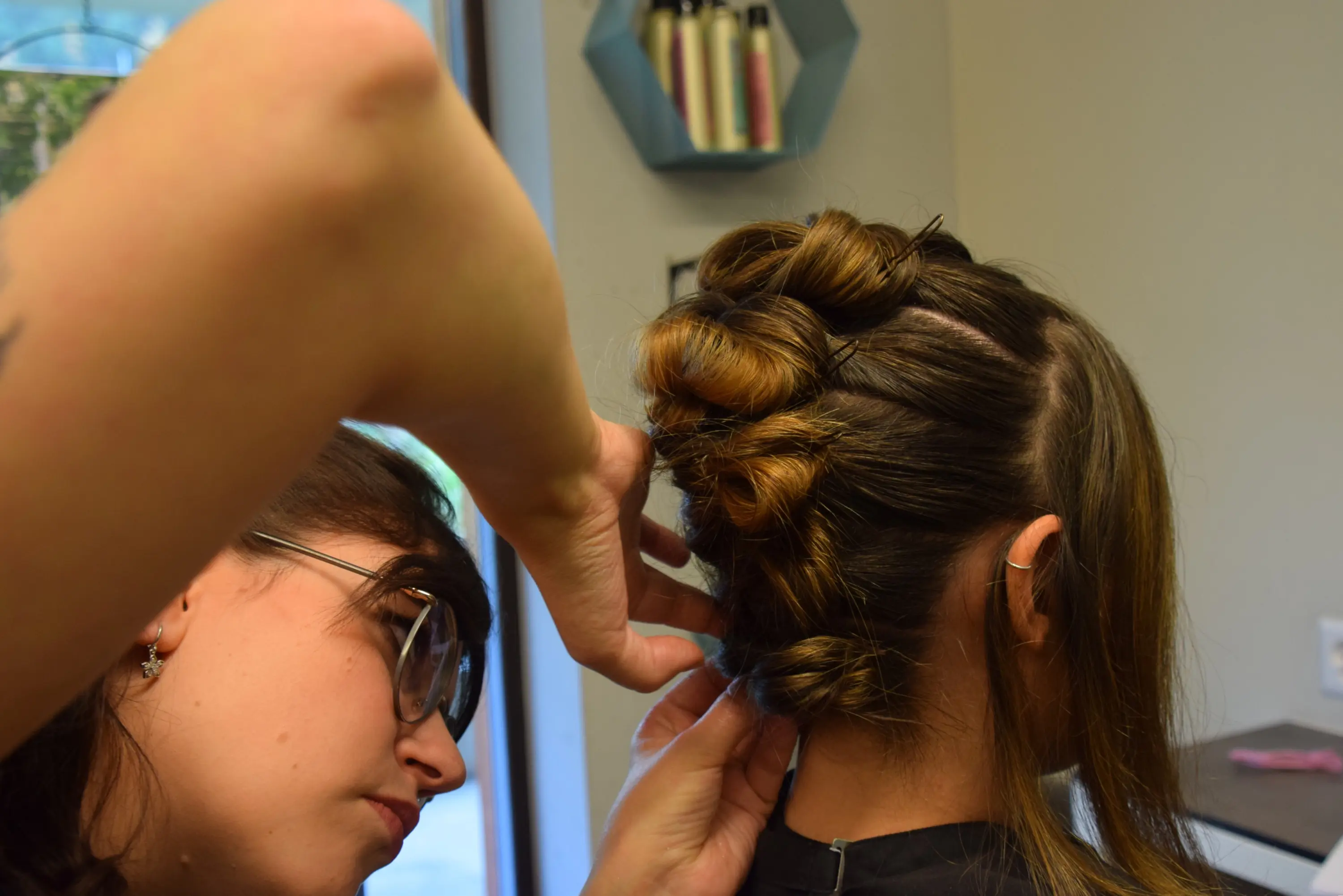 Una parrucchiera sta acconciando i capelli di una cliente, creando un elegante raccolto con trecce. La cliente, seduta, ha i capelli lunghi e lisci.