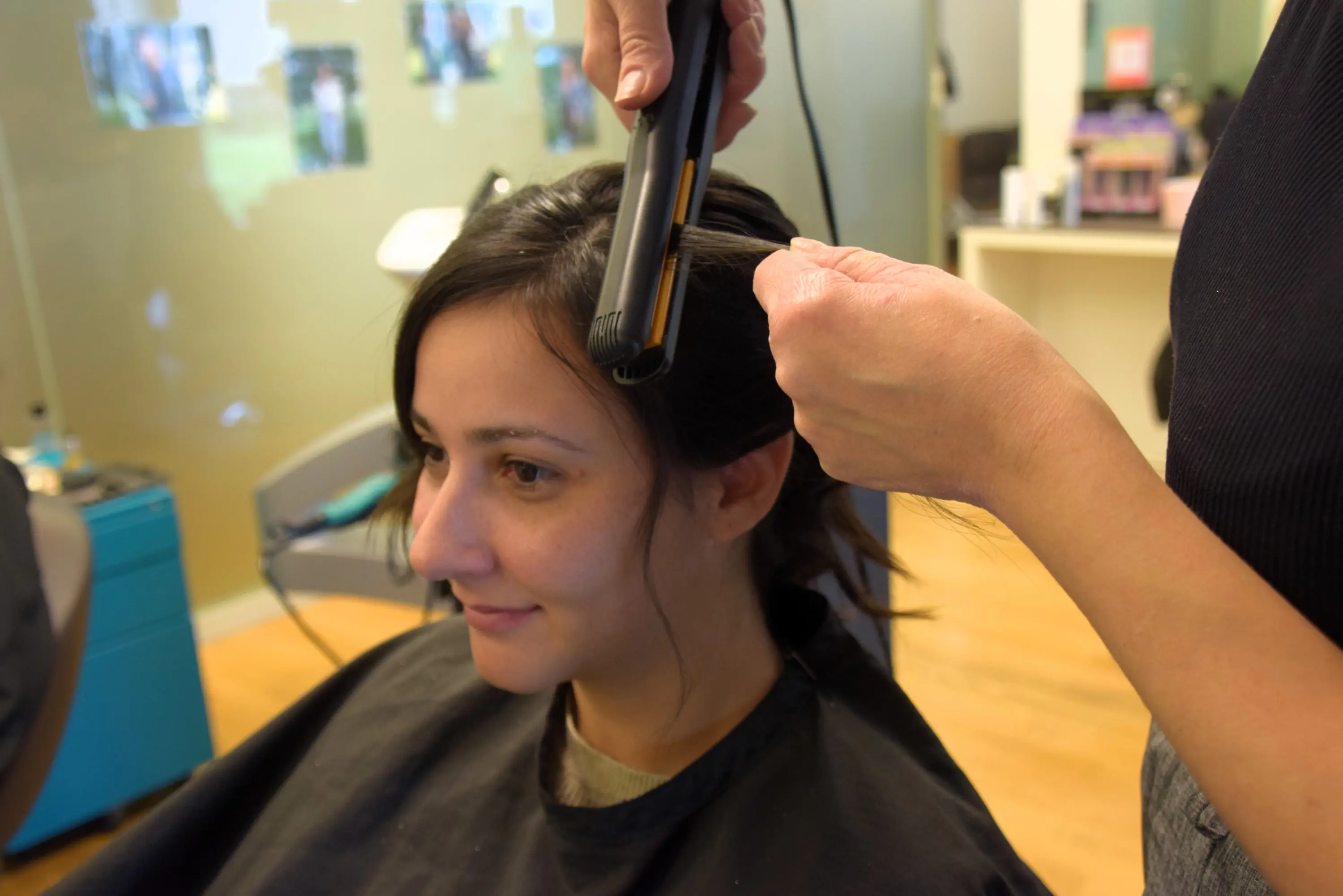 Una donna seduta su una sedia di un salone di bellezza mentre una parrucchiera le liscia i capelli con una piastra.