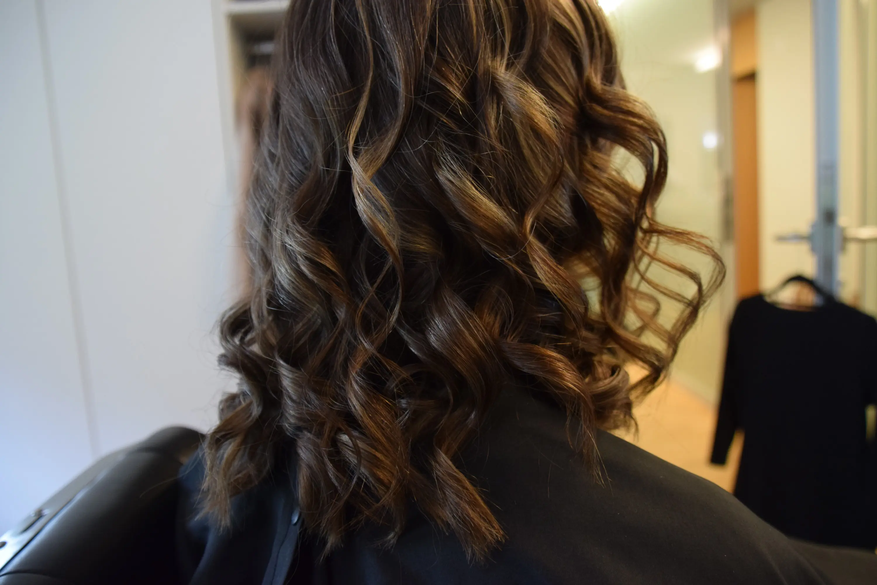 Capelli mossi e ricci di lunghezza media, con riflessi castani, visibile dal retro. Sullo sfondo, una porta e un abito appeso.