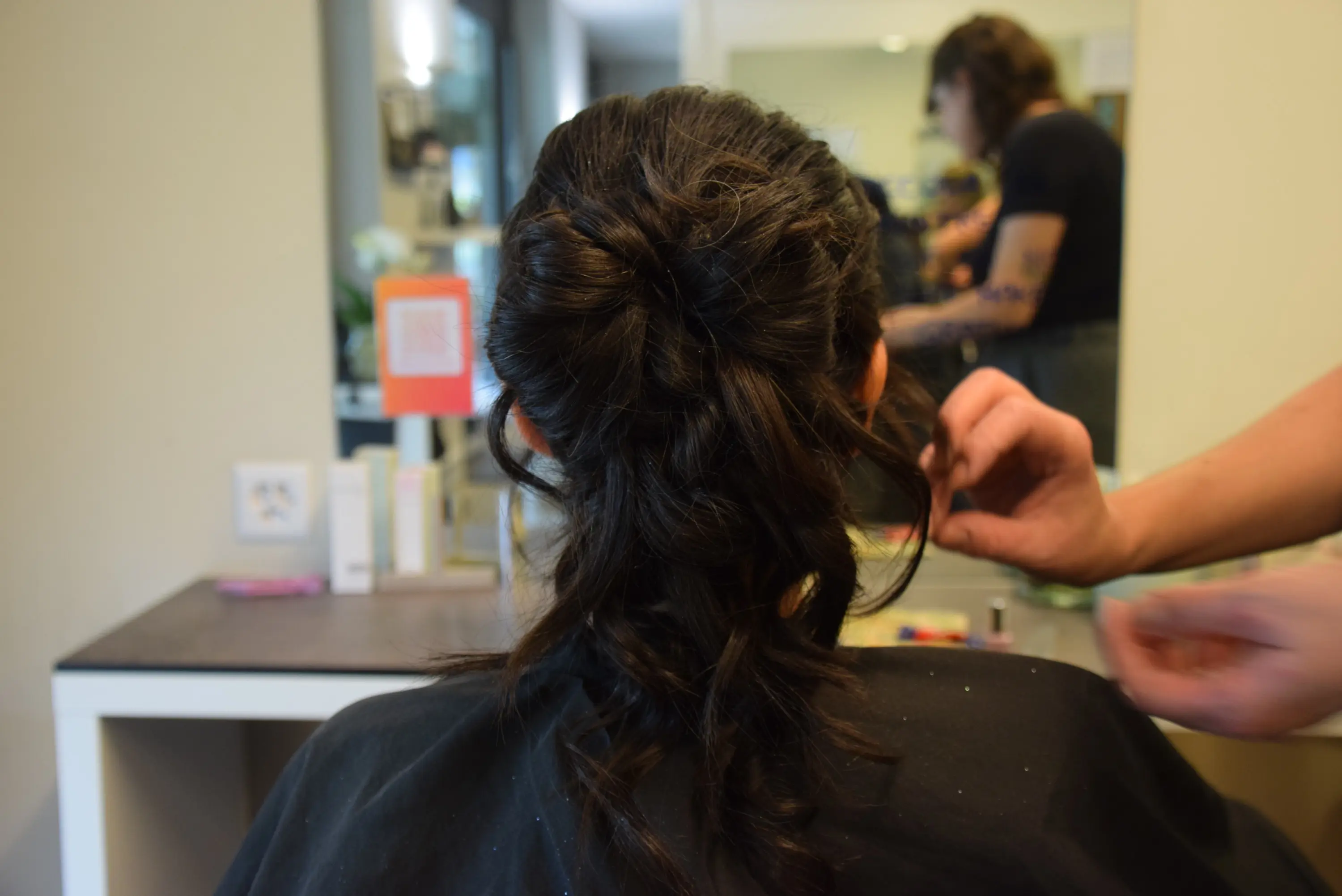 Una persona con capelli scuri in una sala di bellezza, seduta di spalle mentre un parrucchiere sistema i capelli. Sullo sfondo si vede un'altra persona che lavora.