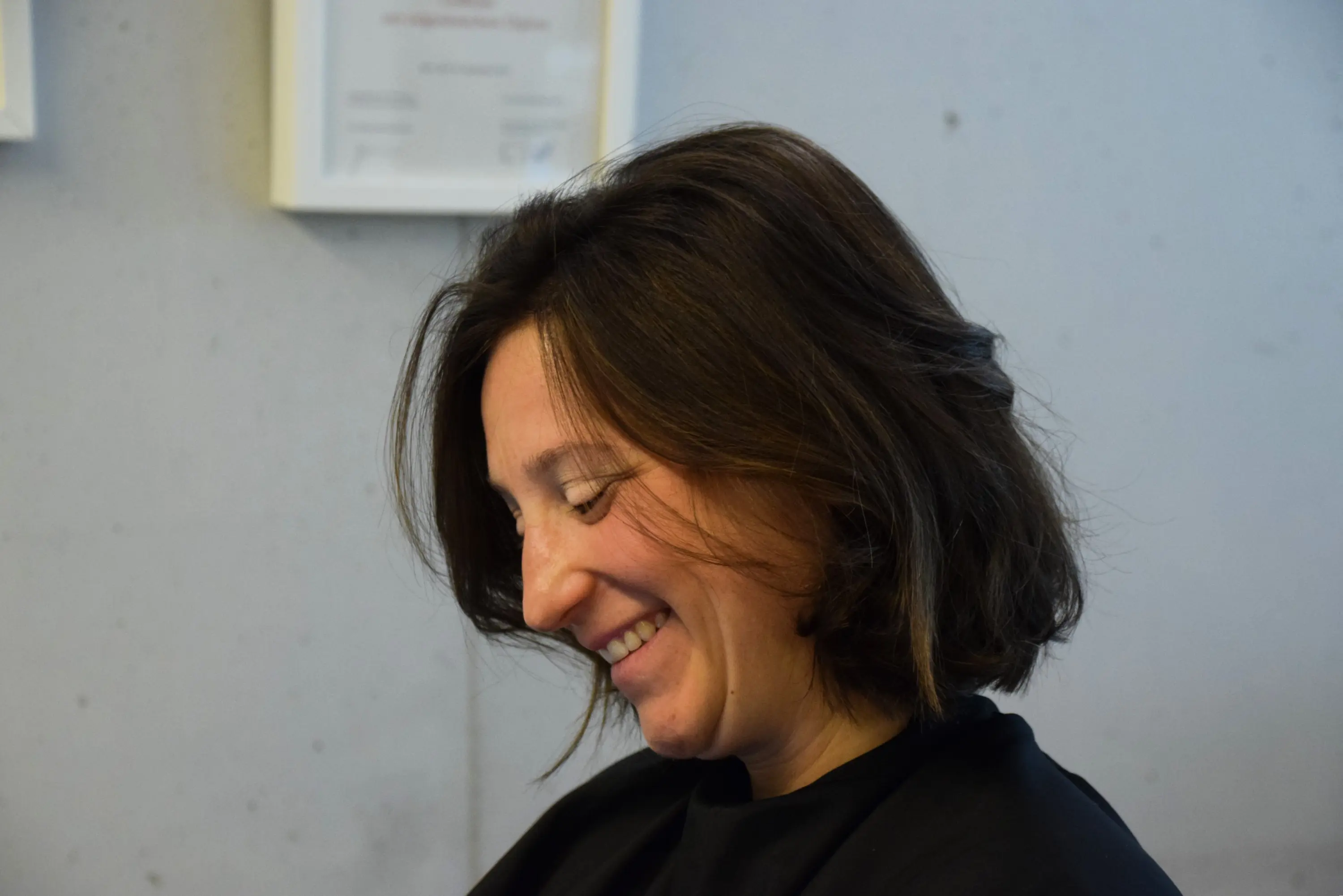 Donna con capelli castani e ondulati, sorridente mentre guarda verso il basso. Si trova in un ambiente moderato con pareti di cemento.