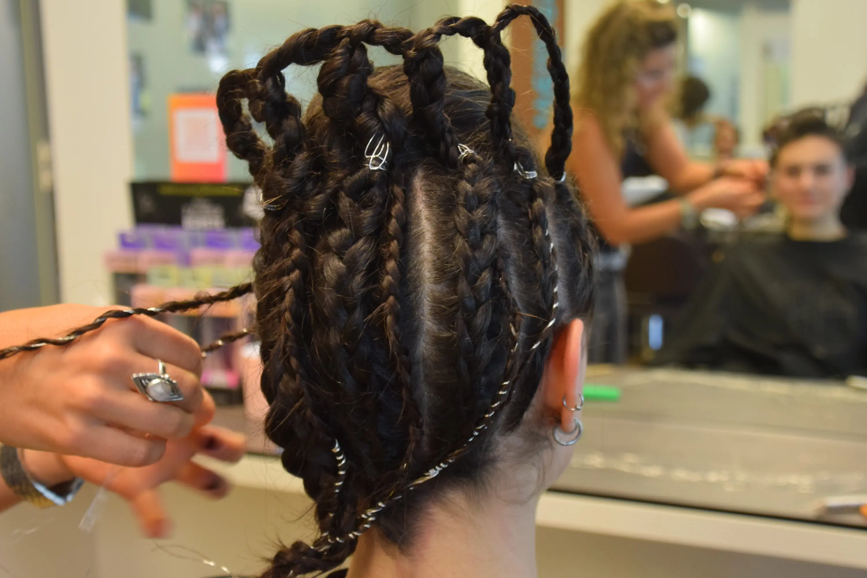 Acconciatura con treccine intricate e clip metalliche. Sullo sfondo si vede un'altra persona seduta al tavolo di lavoro.