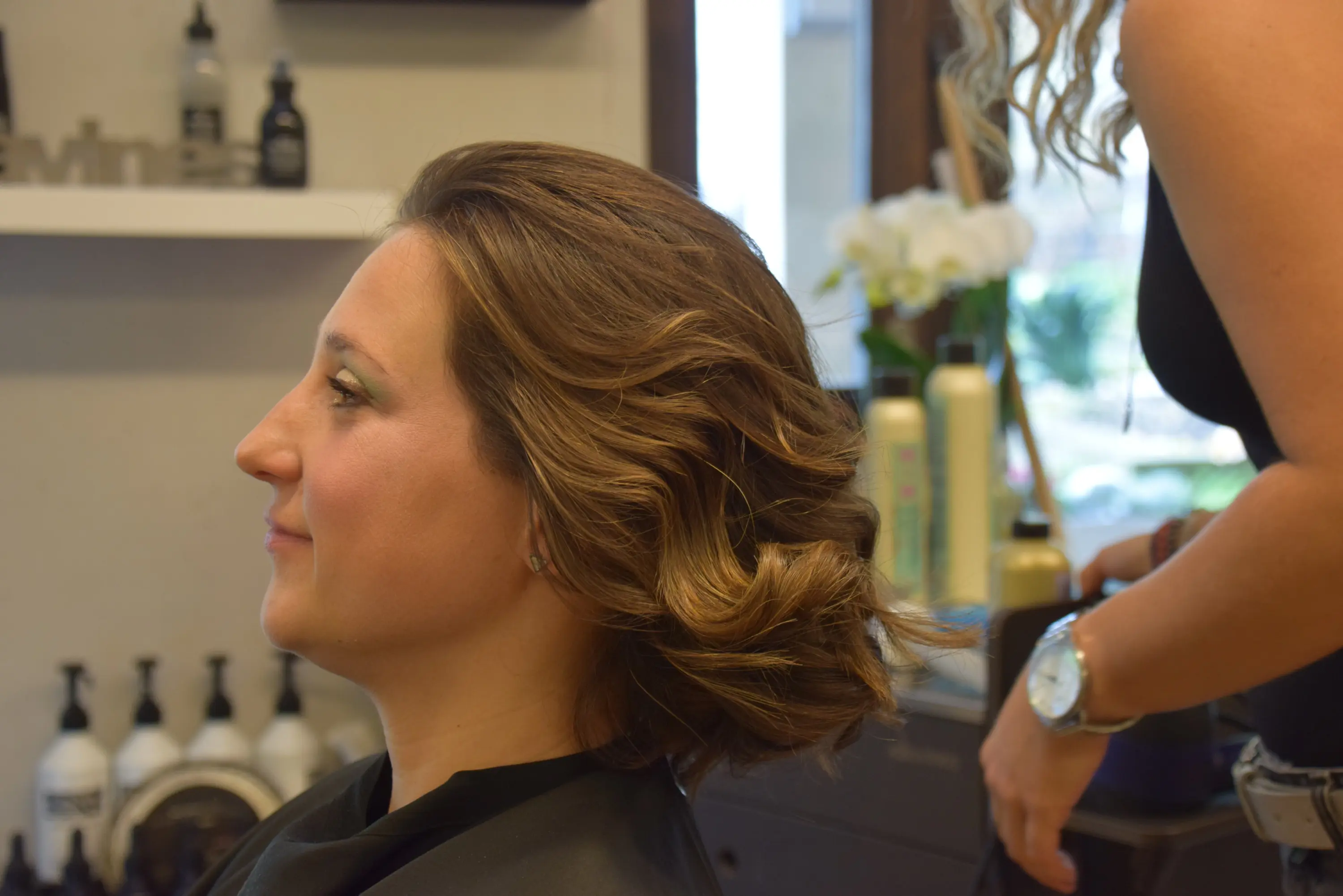 Una donna con capelli mossi e voluminosi è seduta in un salone di bellezza, mentre un'altra persona le sistemano i capelli. Sullo sfondo si vedono prodotti per capelli in scaffali.