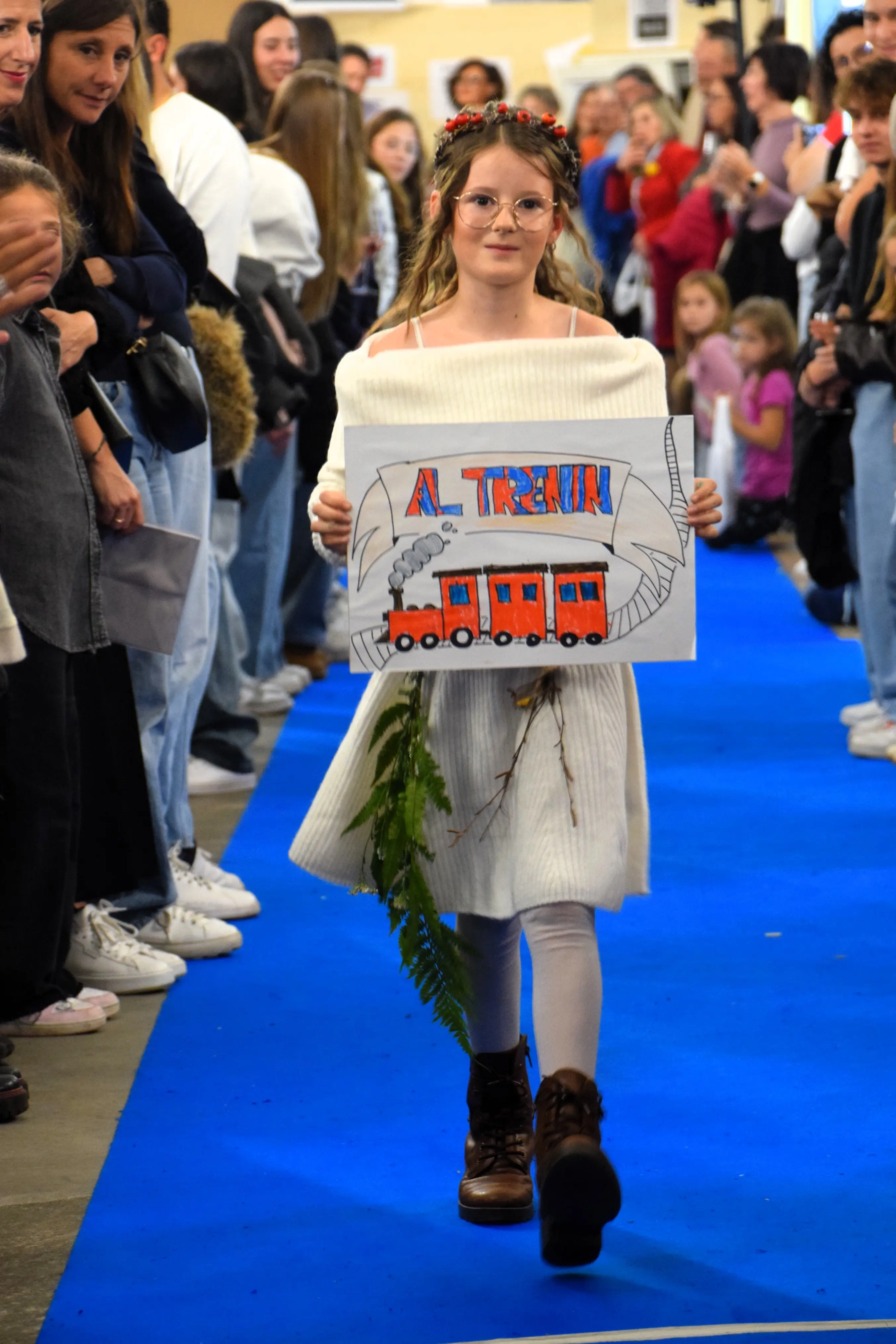 Bambina che sfilano su una passerella blu, indossando un abito bianco e occhiali. Tiene un cartello disegnato con un treno e la scritta "Al treno". Circondata da un pubblico che osserva entusiasta.