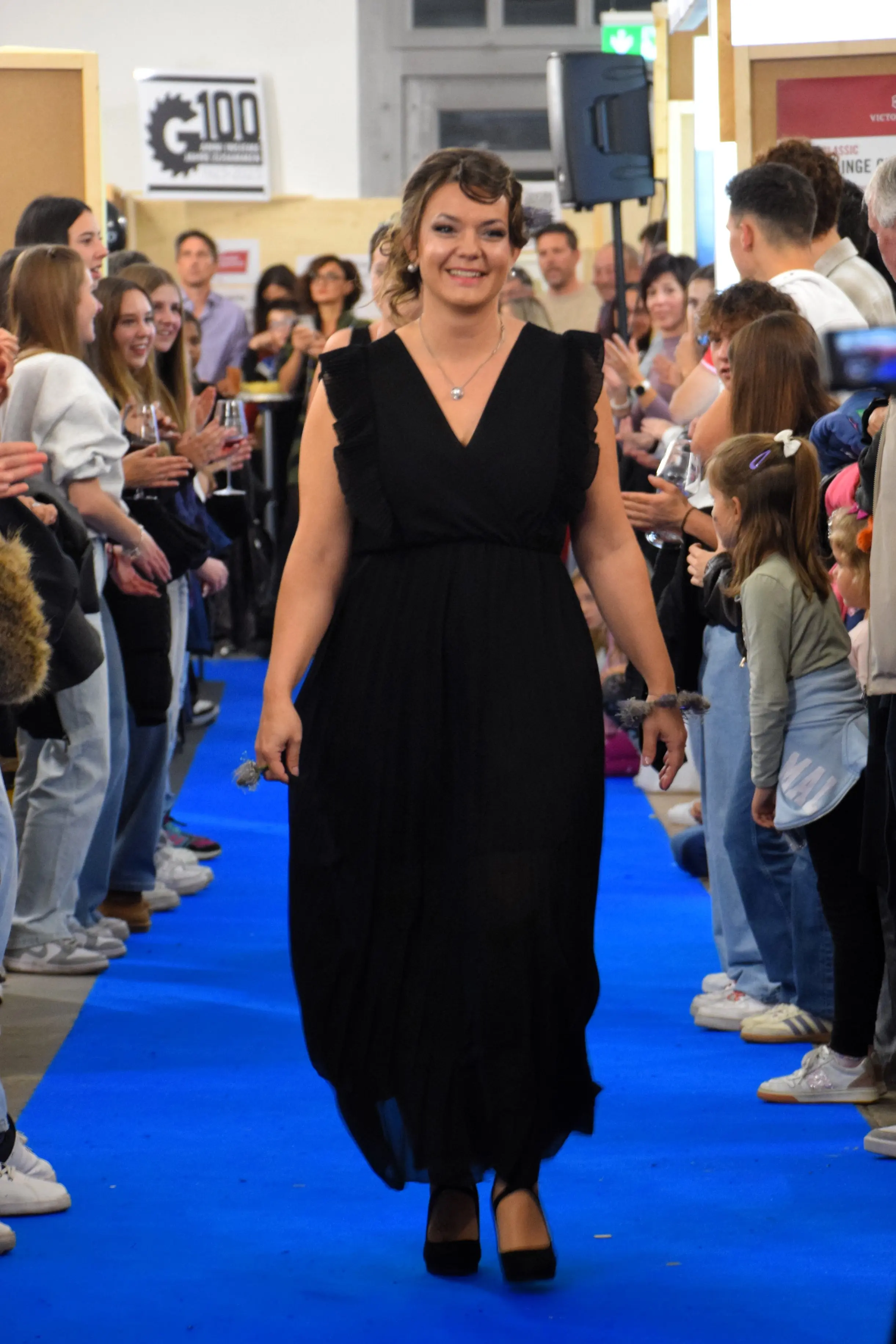 Una donna con un abito nero e capelli ondulati cammina su una passerella blu, sorridendo. Ci sono spettatori che la applaudono ai lati, e si notano anche alcuni bambini.