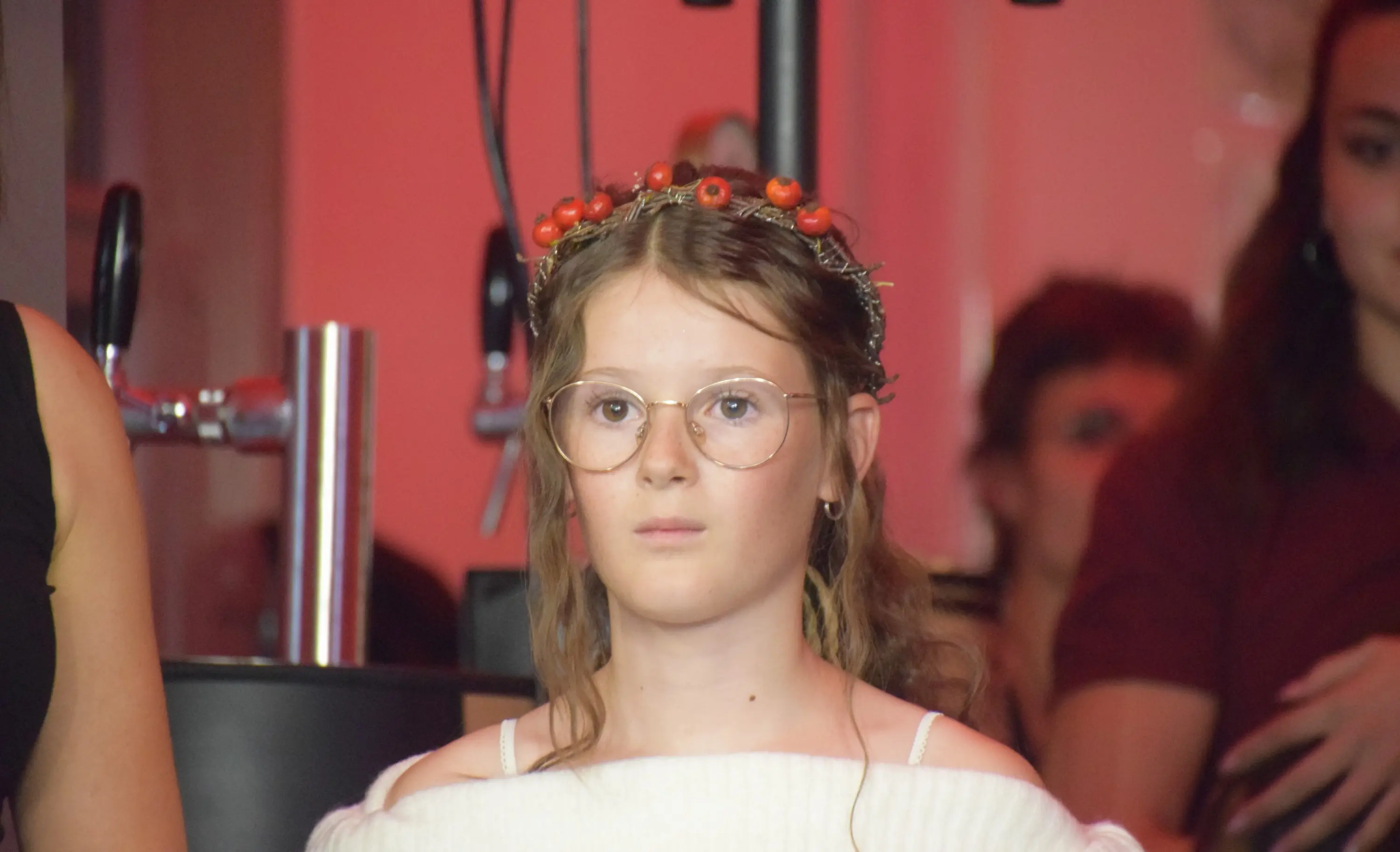 Ragazzina con coroncina di frutti rossi e occhiali, indossa un maglione bianco. Sullo sfondo, altre persone in ombra. Atmosfera calda e luminosa.