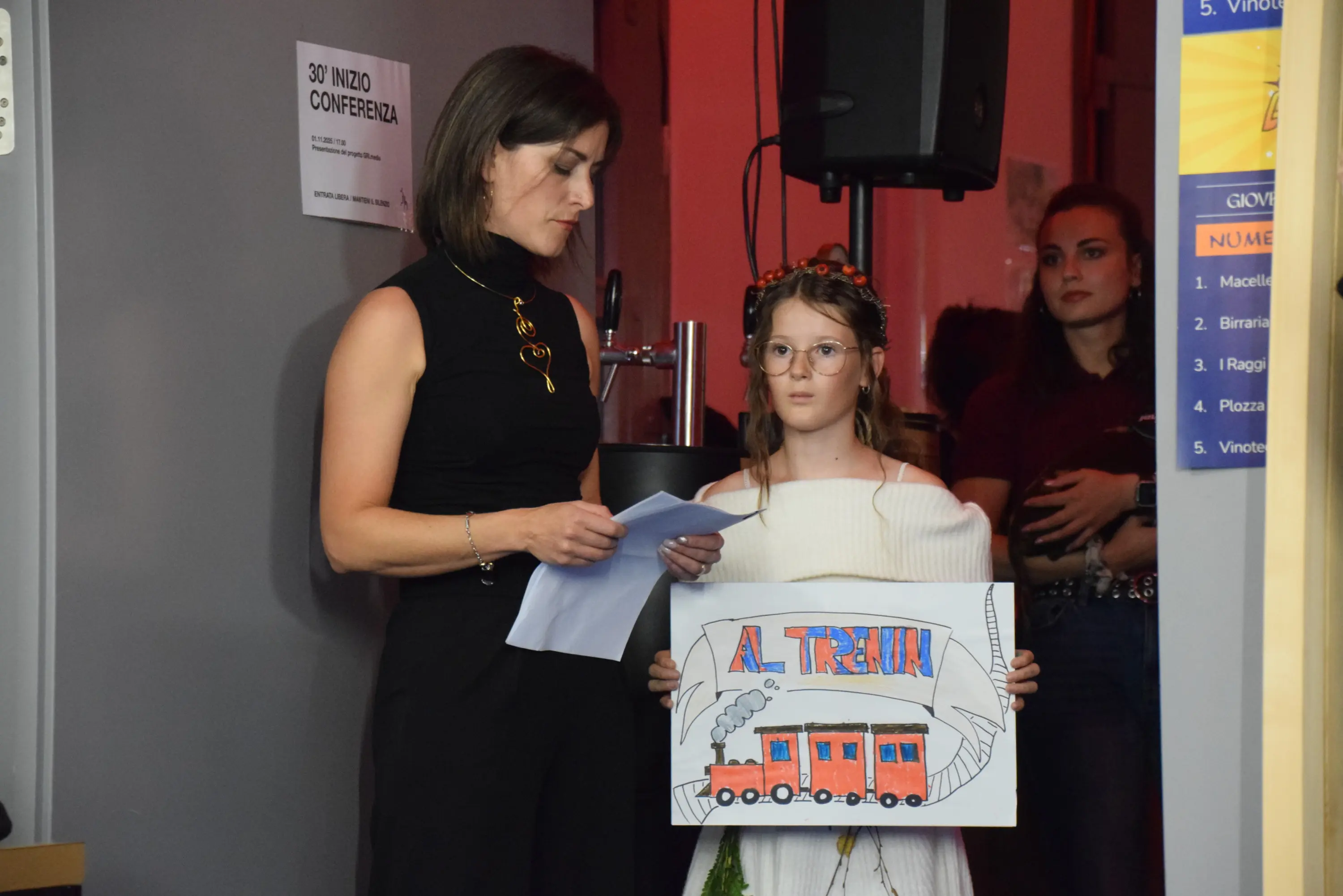 Due donne in un evento, una legge un testo mentre l'altra tiene un cartello disegnato con un treno e la scritta "Al Treno". Sullo sfondo si vedono altre persone e un ambiente moderno.