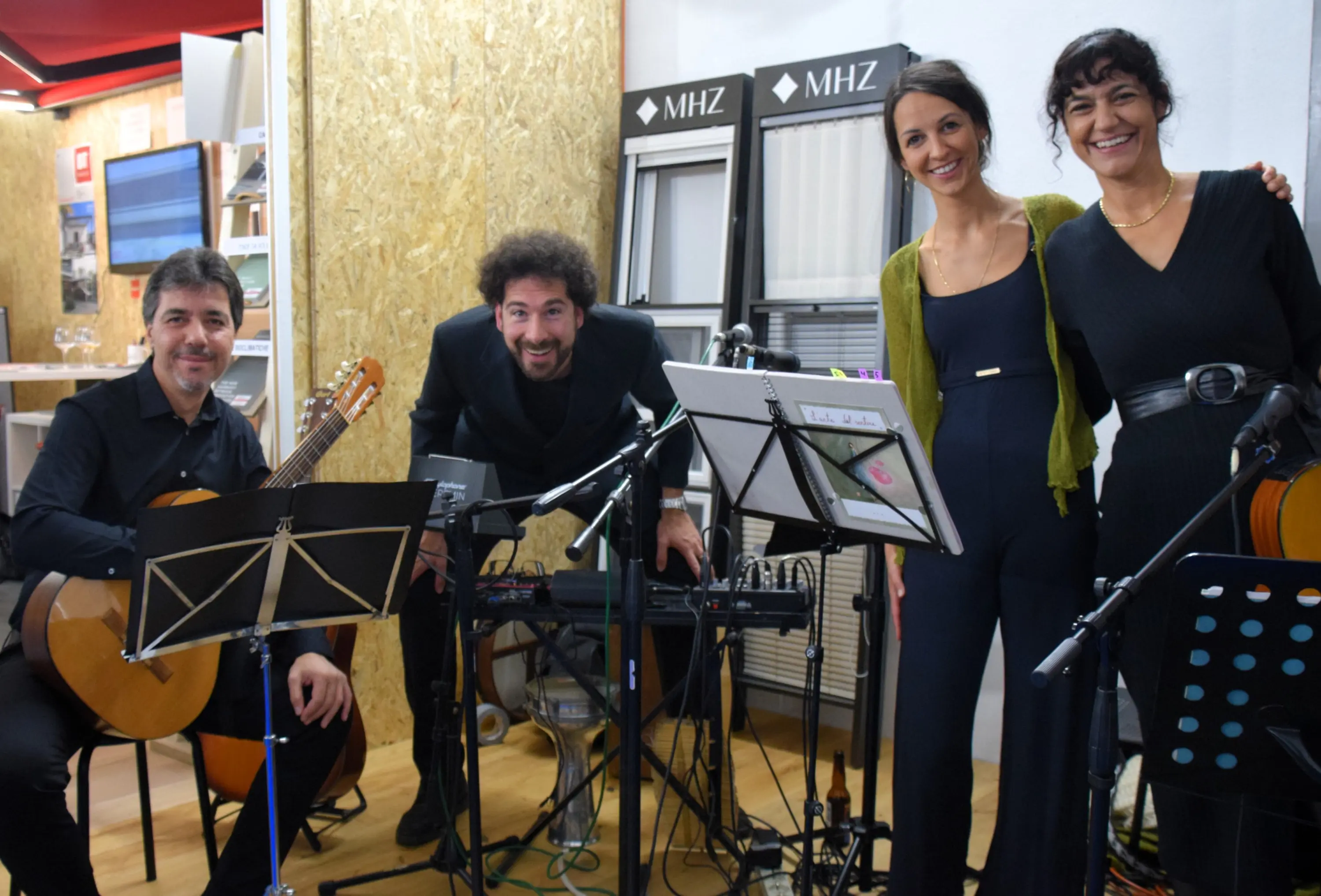 Un gruppo di musicisti sorridenti si esibisce in un locale, con un chitarrista, un uomo al centro con un sintetizzatore, e due donne vestite elegantemente.