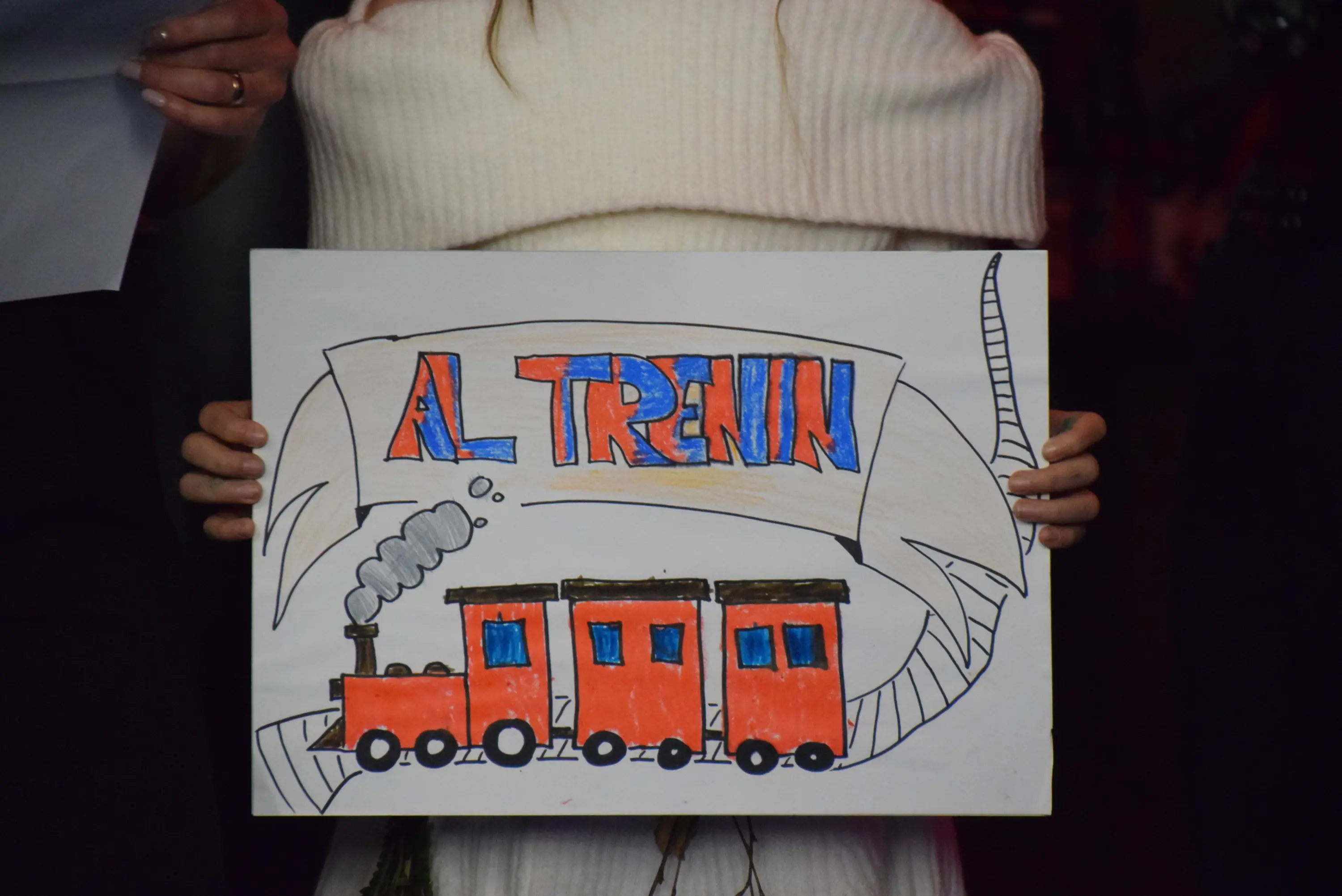Una persona tiene un cartello colorato con la scritta "AL TRENI" e un disegno di un treno rosso.