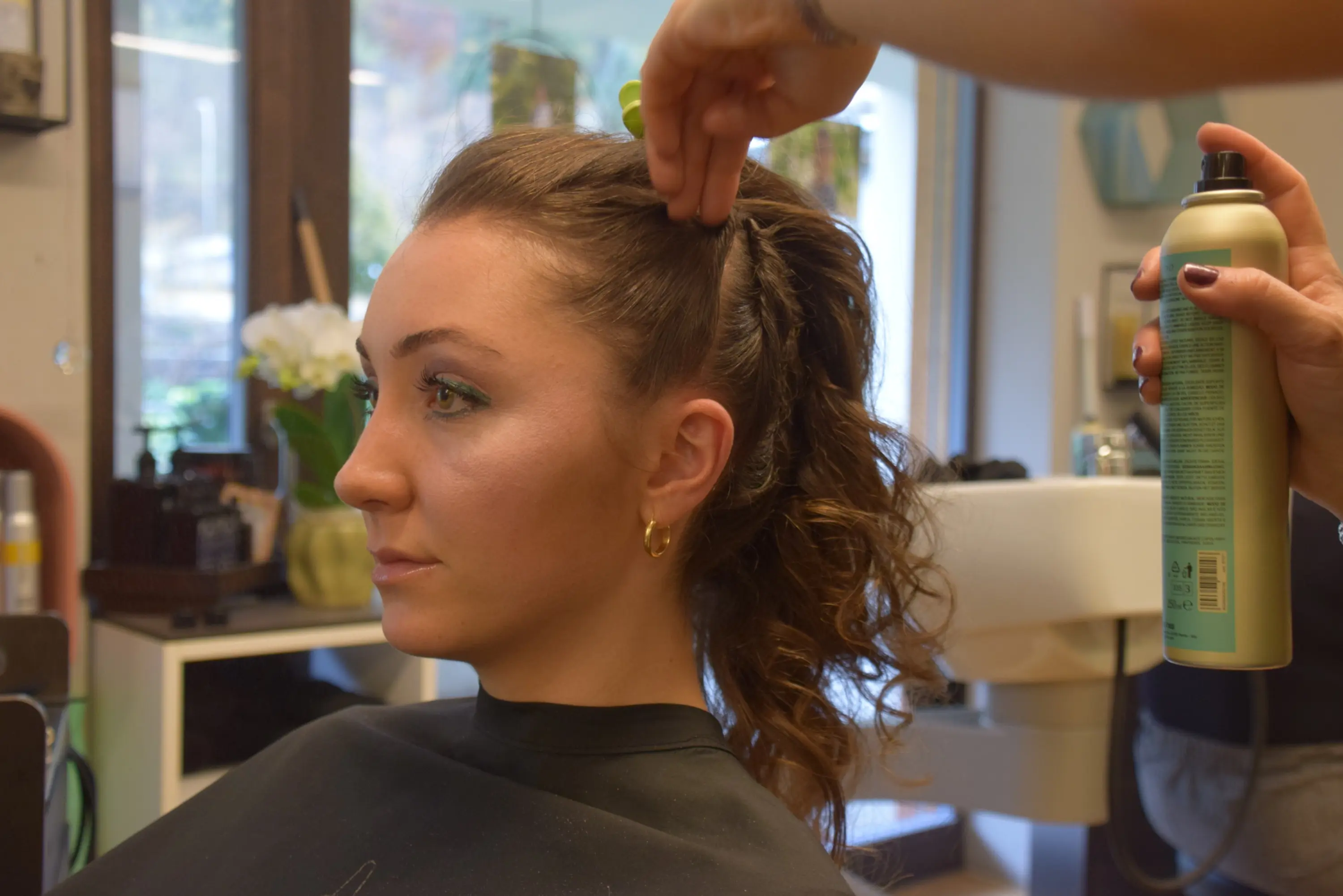 Una donna con capelli ricci e raccolti sta ricevendo un’acconciatura in un salone di bellezza. Un parrucchiere le sistema i capelli mentre tiene in mano una bomboletta di lacca.