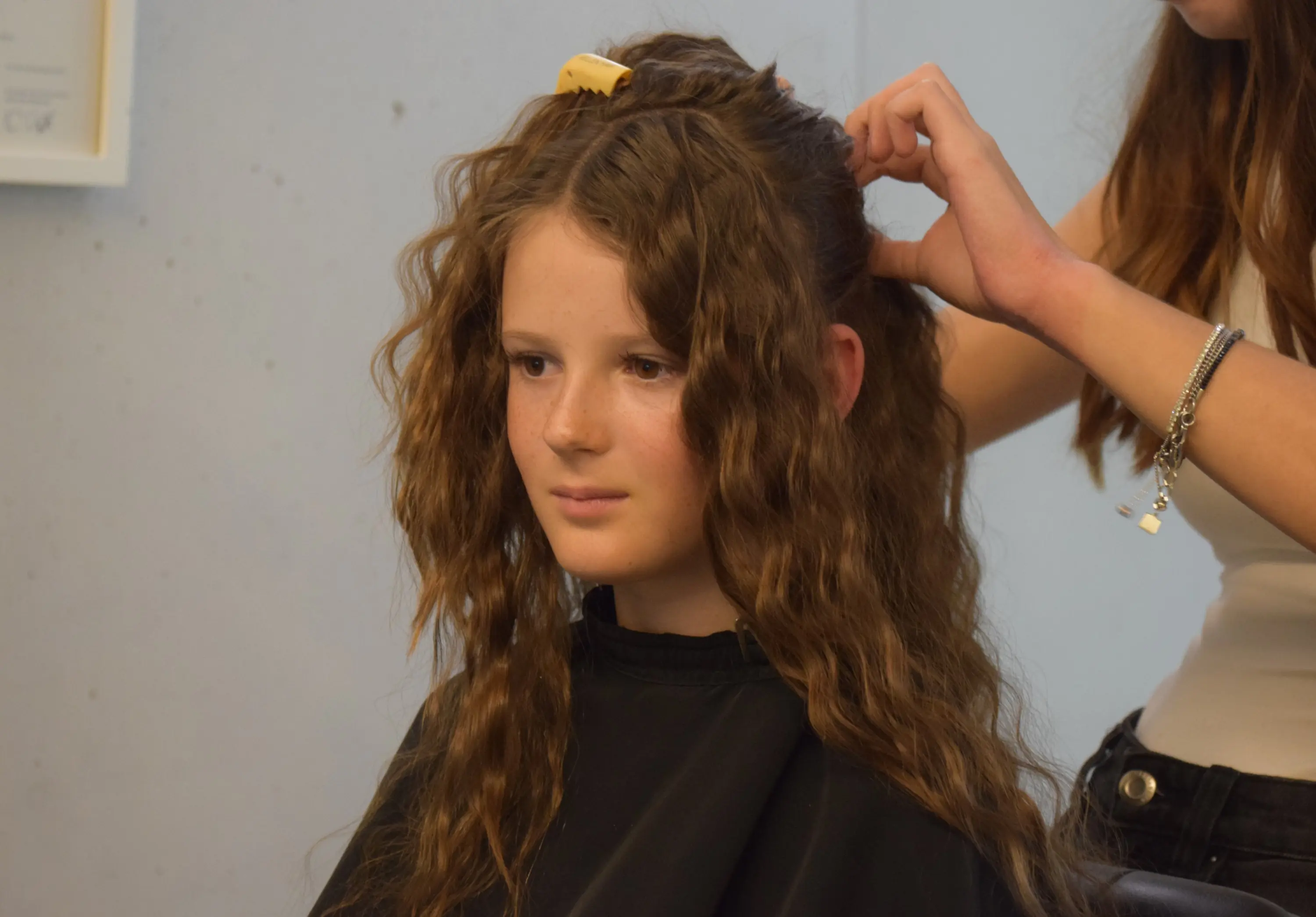 Ragazza con capelli ricci e lunghi seduta mentre una persona le sta sistemando i capelli con una molletta.