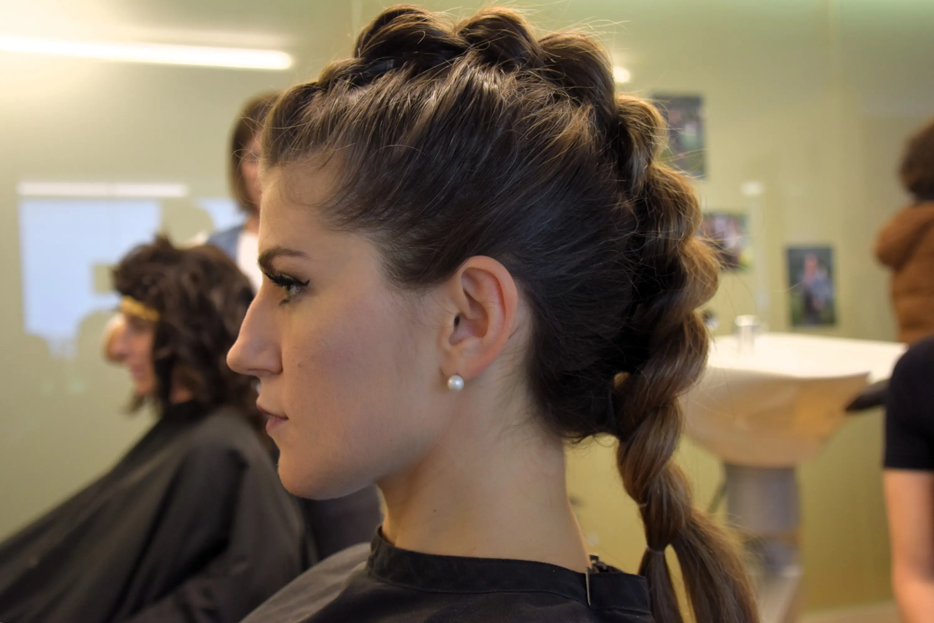 Donna con un'acconciatura a treccia laterale, in profilo. Indossa un orecchino di perla e ha un'espressione seria. Sullo sfondo si intravedono altre persone in un salone di bellezza.