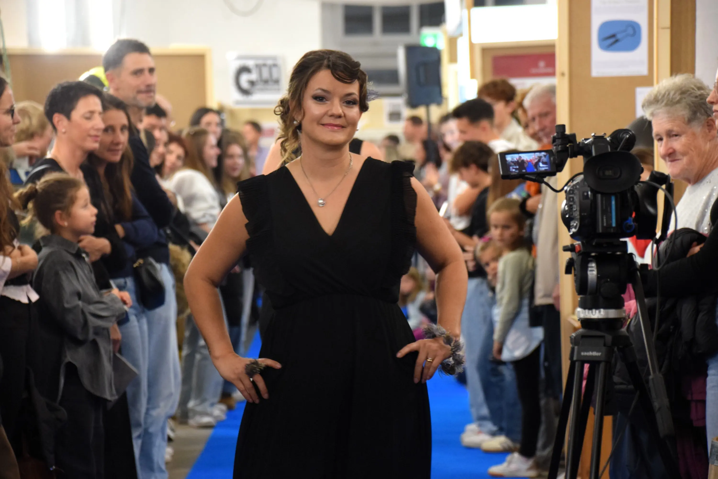 Una donna in abito nero cammina su un tappeto blu durante una sfilata, mentre il pubblico la osserva. Sullo sfondo, si vedono molte persone, alcune delle quali filmando con una videocamera.